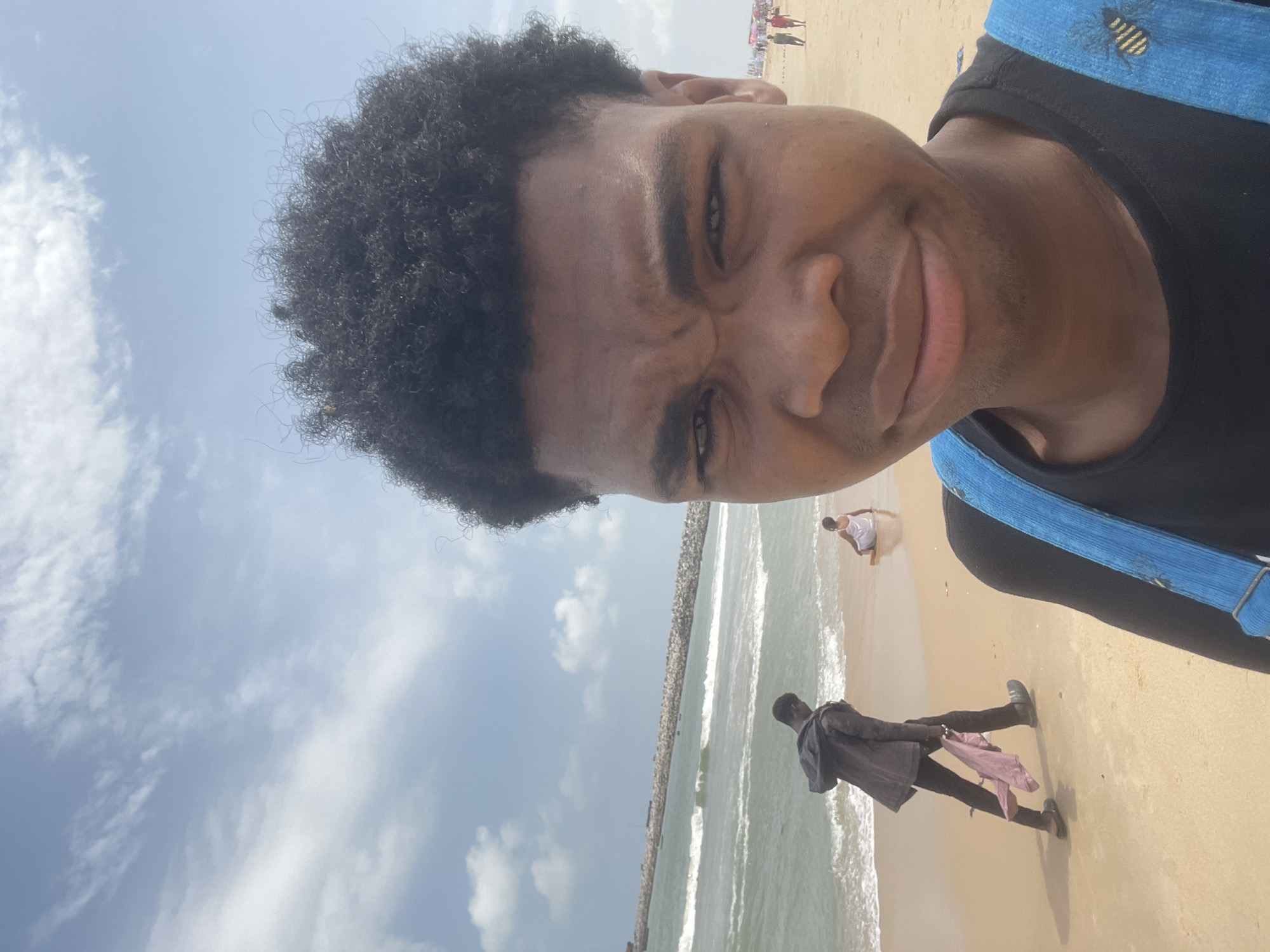A selfie is taken at the beach in Ilado, Nigeria, with a person's face filling the foreground. The person has dark, curly hair and is looking directly at the camera with a slightly distorted expression. They are wearing a black t-shirt with blue straps of a backpack visible. In the background, the sandy beach meets the ocean under a cloudy sky. To the left, a person in a dark jacket and black pants walks along the shoreline, carrying a light-colored cloth. Further down the beach, a person in a white garment is sitting. In the distance, a breakwater juts into the sea, and a few more people are visible near beach umbrellas.