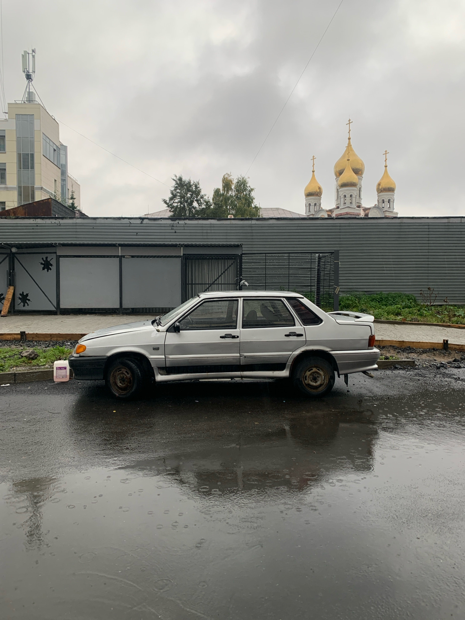 An overcast day in Arkhangelsk, Russia. A silver sedan is parked on a wet asphalt road reflecting the cloudy sky. The car, showing signs of wear, has a slightly ajar hood. To the left of the car, a white plastic container with pink liquid sits on the roadside. Behind the car, a grey metal fence stretches horizontally across the scene. Beyond the fence, a building with a rusted roof is visible, and further in the background, the golden domes of a church rise above the rooftops. The scene is characterized by a general atmosphere of dampness and muted colors, typical of rainy weather.