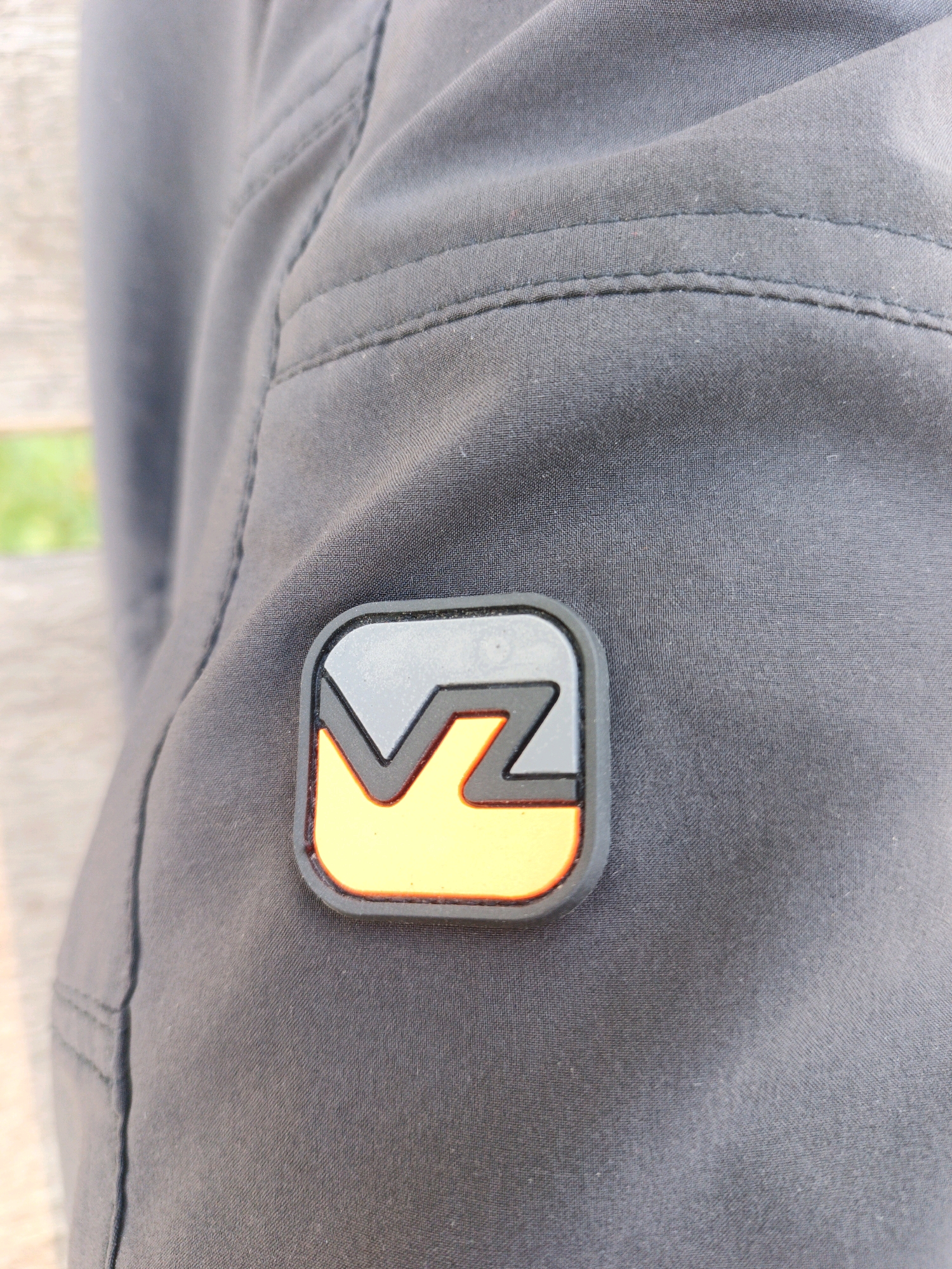 The image shows a close-up view of dark gray fabric with stitching, likely from a pair of pants or shorts. A prominent logo patch is affixed to the fabric. The patch is square-shaped with rounded corners and features a design in shades of gray and yellow, outlined in black and orange. The gray portion of the logo has a stylized "VZ" design. The background is blurred, suggesting an outdoor setting with hints of greenery and a wooden surface. The location is Unknown, Unknown.
