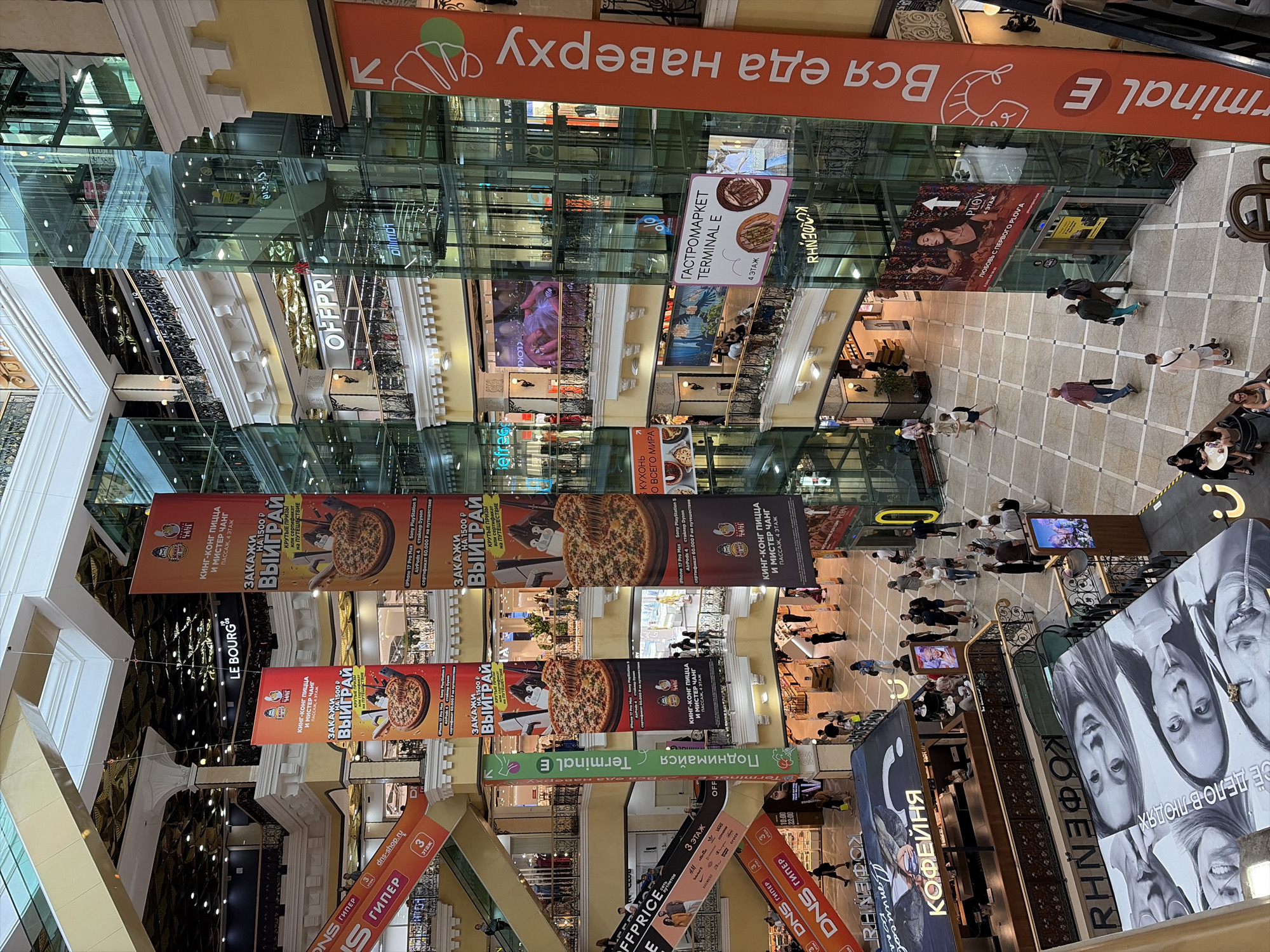 The image shows a high-angle interior view of a multi-story shopping mall in Yekaterinburg, Russia. The mall features a large atrium with glass elevators and escalators connecting different levels. Various storefronts and advertising banners are visible throughout.
In the foreground, a large banner displays black and white portraits of people, likely for an advertisement. Below this, a cafe or food stall labeled "КОФЕЙНЯ" is visible. Escalators are also prominent, with orange banners displaying "DNS ГИПЕР" advertising an electronics store.
Several large vertical banners hang from the ceiling, advertising "KING KONG PIZZA" and offering prizes. Other banners advertise "OFFPRICE" and "GASTRONOMARKET TERMINAL E". An orange banner on the right side of the image has large vertical text in Russian, "Вся еда наверху", translating to "All food upstairs".
People are seen walking, shopping, and interacting within the mall environment across various levels. The architecture appears modern, with extensive use of glass and light-colored materials. The overall scene depicts a bustling commercial space within a Russian city.
