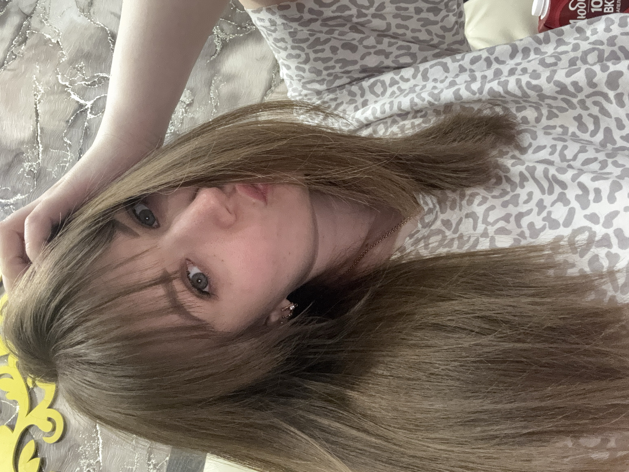 The image captures a young woman lying down or reclining indoors, looking directly at the camera with a neutral to slightly pensive expression. Her head is tilted, and her long, light brown hair, possibly with bangs, cascades around her face and across the surface she's resting on. Her left eye, which appears blue or grey, is visible, along with her nose and mouth, where her lips are slightly parted. There are visible freckles on her nose and cheek. Her left arm is bent, with her hand resting near her head.
The setting appears to be a casual indoor space, likely a bedroom or living room. The woman is resting on what seems to be a bed or couch covered with light grey and white leopard-print fabric or bedding. A decorative yellow-gold element, possibly part of a wall decoration or furniture, is visible in the lower left corner. In the upper right background, a red can is partially visible. Cyrillic text on the can includes "Люби" (meaning "Love"), "10", "ВКУ" (likely part of "вкус" meaning "taste"), and "АПЕЛ" (short for "апельсин" meaning "orange"), suggesting it is a can of orange-flavored beverage. The lighting is soft and even, indicating an indoor shot during the day or in a well-lit environment.