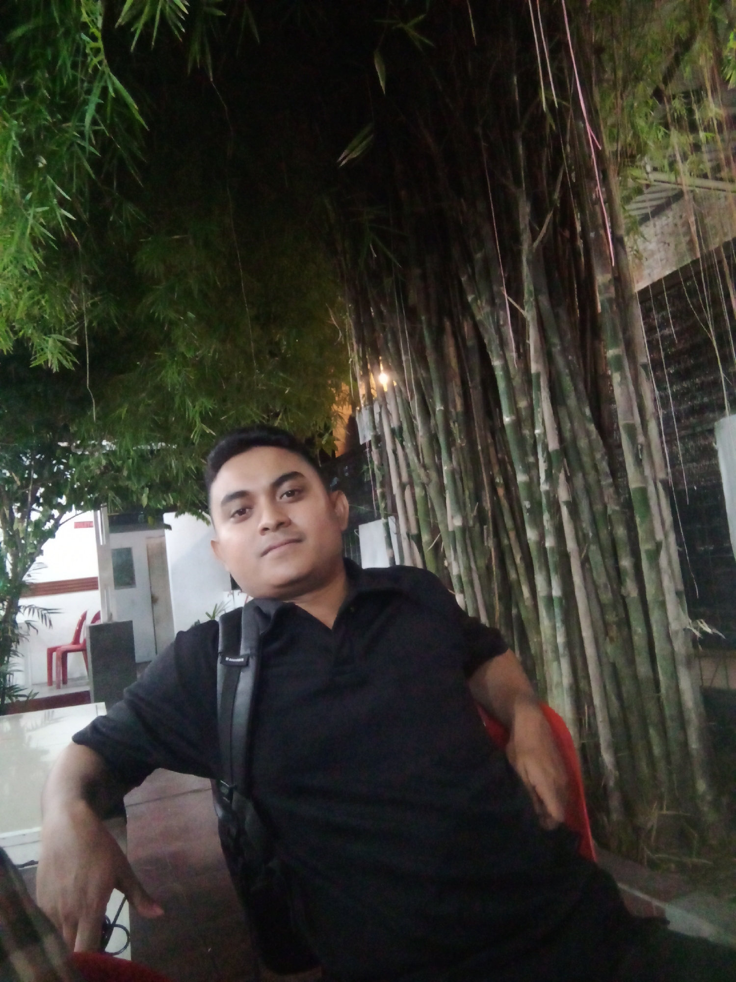 The image shows a man seated outdoors, likely in a public or semi-public space. He is wearing a black polo shirt and has a black bag slung over his shoulder. The background features dense bamboo stalks and foliage. To the left of the frame, a building is visible, and a red plastic chair. The location is most likely somewhere in the city of Lhokseumawe, Indonesia.