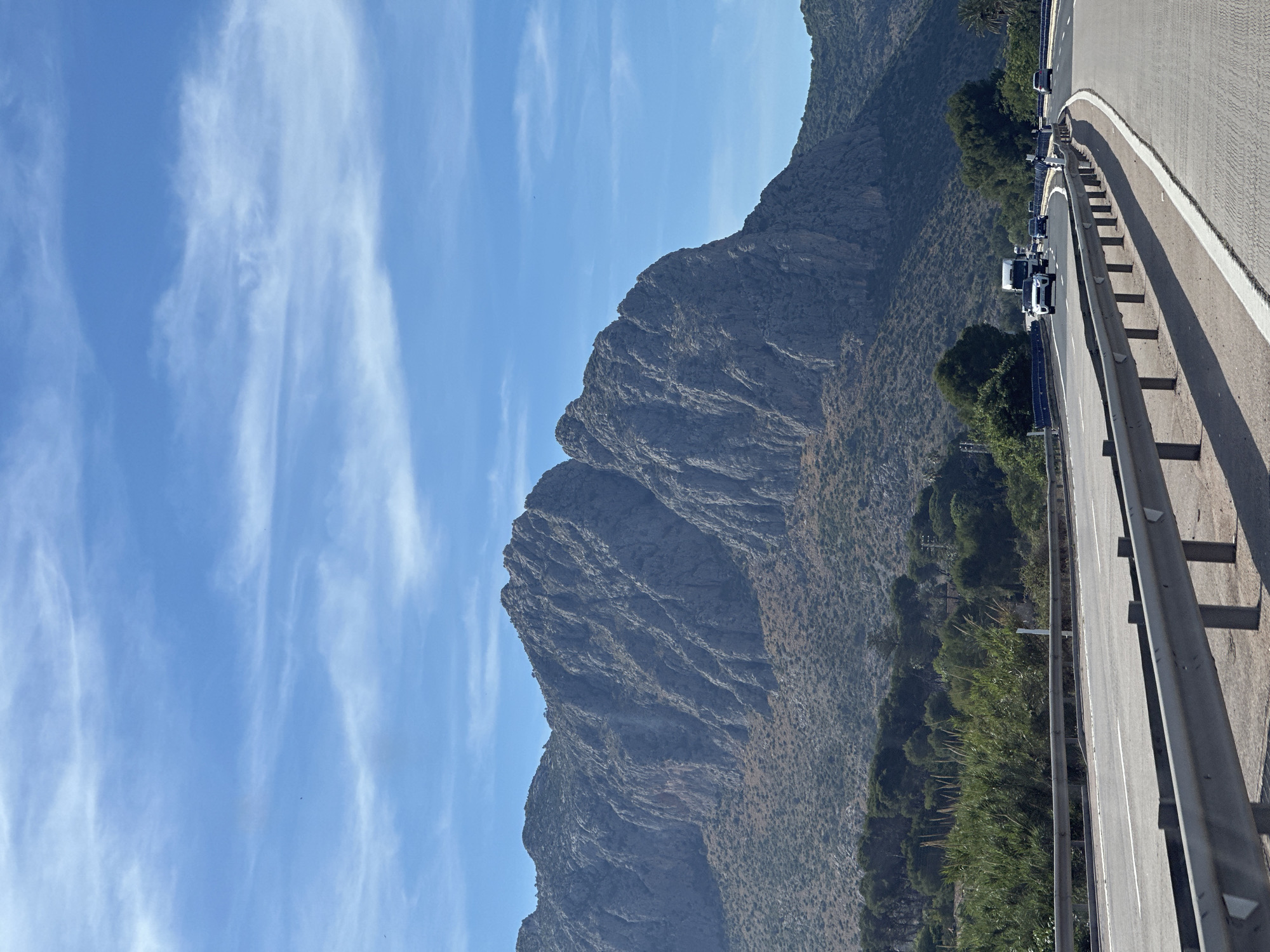The image shows a view from a highway in Ondara, Spain, featuring a mountainous landscape under a blue sky with wispy clouds. The road, seen on the right, has a metal guardrail and light-colored asphalt with white lane markings. Traffic is backed up on the highway. A large mountain is centered, covered in rocky terrain and sparse vegetation.