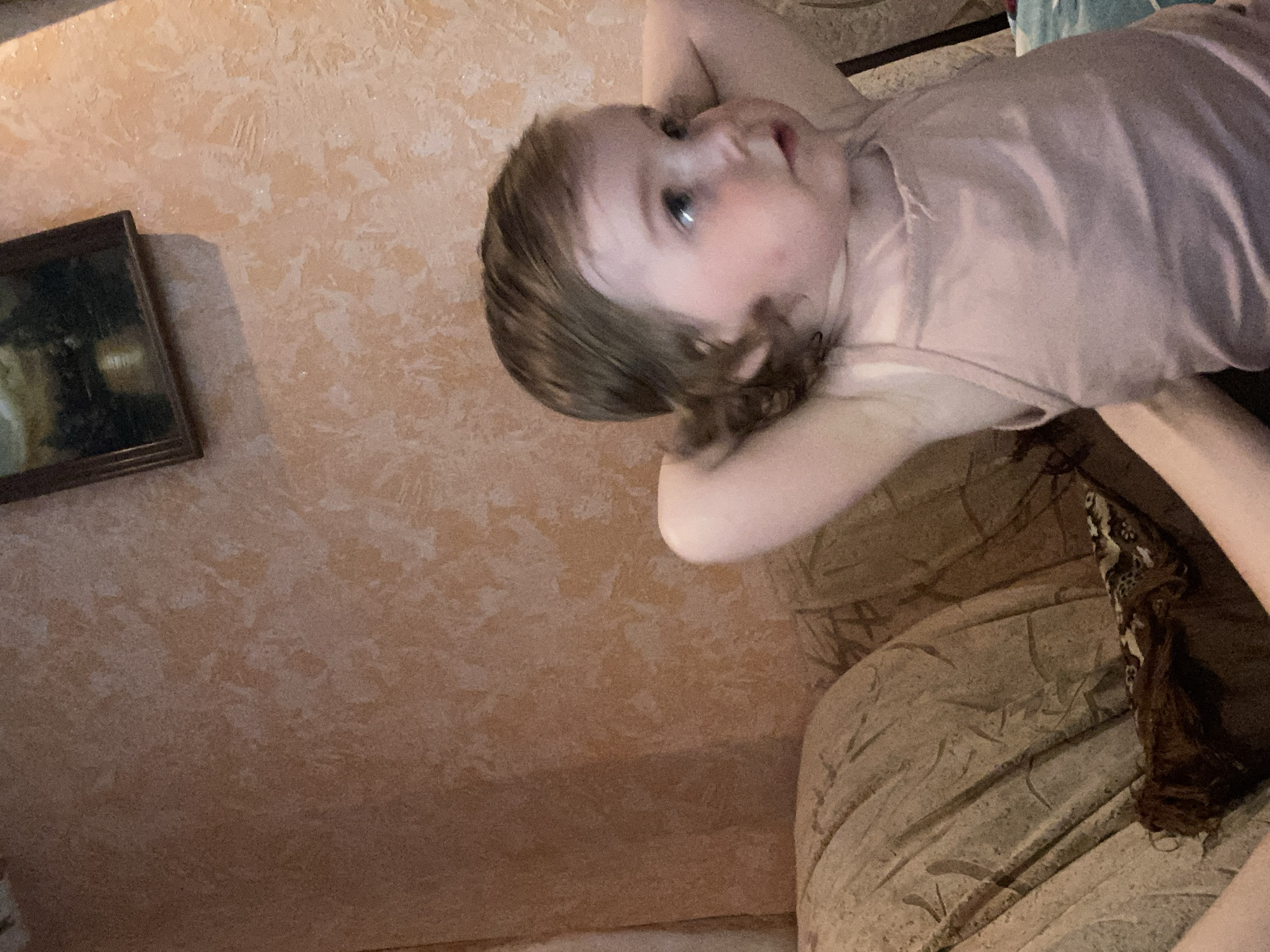This is a vertical, close-up, indoor photograph of a baby lying on their back. The baby, who appears to be very young, has light skin and light brown hair. They are looking upwards and to the right, with a soft, curious expression. The baby is wearing a light pink t-shirt.
The background consists of a textured, peach-colored wall. To the left of the baby, a framed picture hangs on the wall. The frame appears dark and ornate, and the picture within is dimly lit and difficult to discern. Below and to the right of the baby, a portion of a couch or upholstered furniture is visible. The fabric of the furniture has a textured pattern in shades of brown and beige.
The lighting in the room appears to be artificial and somewhat dim, casting shadows and creating a warm, intimate atmosphere. The focus is on the baby's face, highlighting their features and expression. The overall scene suggests a relaxed, domestic setting, likely a living room or bedroom. There is no visible text in the image. The time of day and weather conditions are not discernible from the photograph.