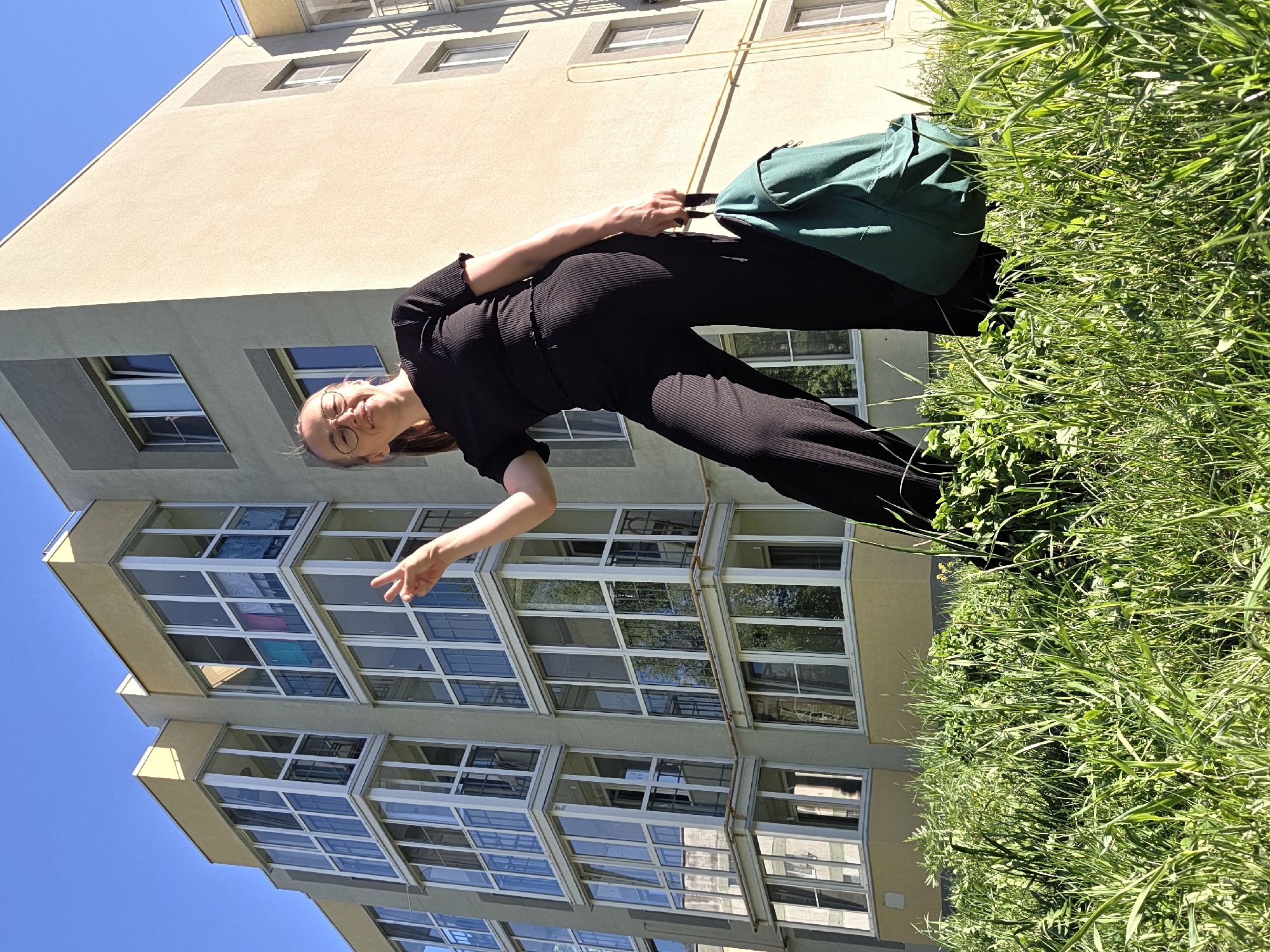 The image depicts a woman standing in a grassy area next to a building. The building is light beige and gray, with many windows that reflect the sky. Some windows have laundry hanging in them. The sky is blue. The woman is wearing a black jumpsuit and glasses and is holding a green backpack. Her right hand is raised, making a peace sign. The grass is tall and green, indicating a park or undeveloped area in an urban setting. There are no readily discernible architectural details or signage to specifically locate the scene in Chaikovsky, Russia.