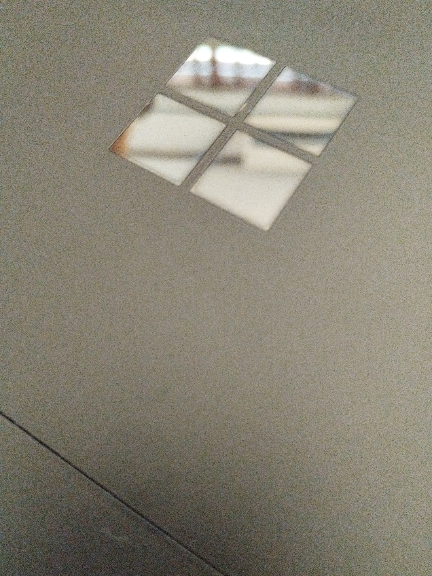 The image shows a close-up, eye-level, outdoor shot of a segment of a grey surface with a pattern of four diamond-shaped reflective elements arranged in a square formation. The reflective elements are angled and appear to be made of mirror or polished metal. They reflect light and the surroundings, showing blurred shapes and colors, possibly indicating an indoor environment with artificial lighting or an overcast sky. The surface itself is matte and textured. There is a faint diagonal line or seam visible across the lower left portion of the image. There are no visible people, animals, or vegetation. The image does not contain any specific landmarks or clues that would allow for its location to be identified within the city of Bogor, Indonesia.