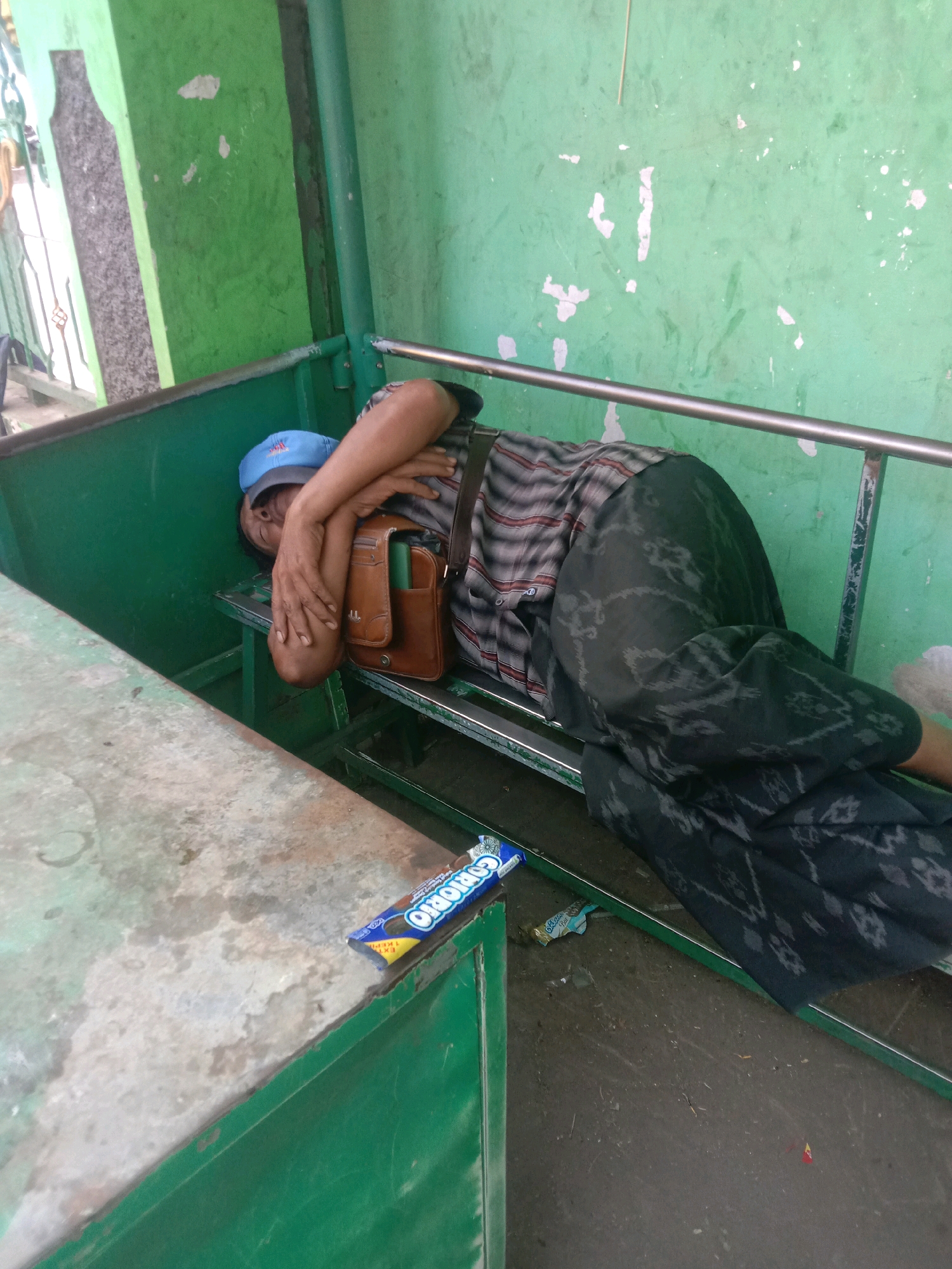 A man wearing a blue cap and a plaid shirt is sleeping on a green metal bench. He has a brown leather bag slung across his chest and is covered with a patterned sarong. A chocolate bar wrapper is visible on the bench in front of him. The background consists of a faded green wall with peeling paint and a metal railing. The scene appears to be outdoors, possibly in a public waiting area, in Karanganyar, Indonesia.