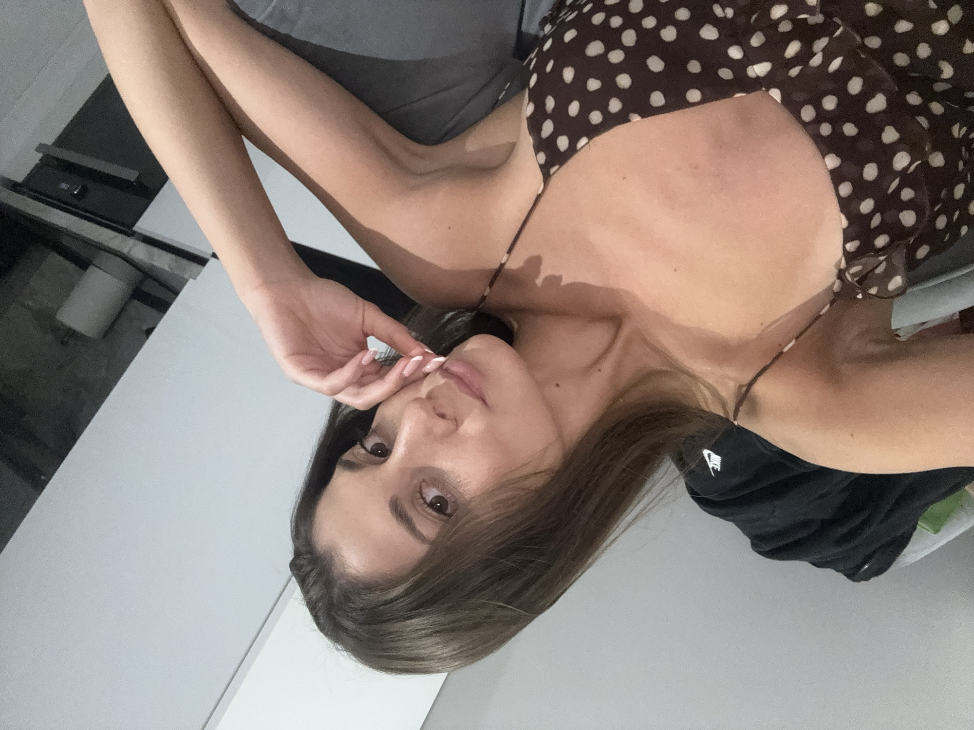 A woman is lying on a bed. She has long brown hair and is wearing a brown and white polka dot dress. She is touching her face with her left hand. The background is a mix of gray and white, appearing to be an indoor space, with a hint of black appliances visible. It is not possible to determine the location in Burgas, Bulgaria from the image.