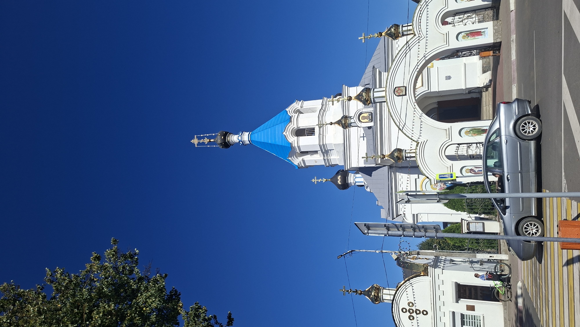 A bright, clear day with a deep blue sky. In the background, a white church with a vibrant blue dome and gold crosses is prominent. Several smaller gold-domed structures and arched entrances with religious iconography are also visible. A mature tree with green foliage is in the upper left corner of the frame. In the foreground, a silver sedan is driving on a paved road. To the left of the road, a pedestrian walkway with yellow and white stripes is visible, and a person with a bicycle is standing near the church entrance. Traffic signals and a street sign are positioned between the road and the church. The scene appears to be in the city of Pastavy, Belarus, given the architectural style of the church and the overall setting.