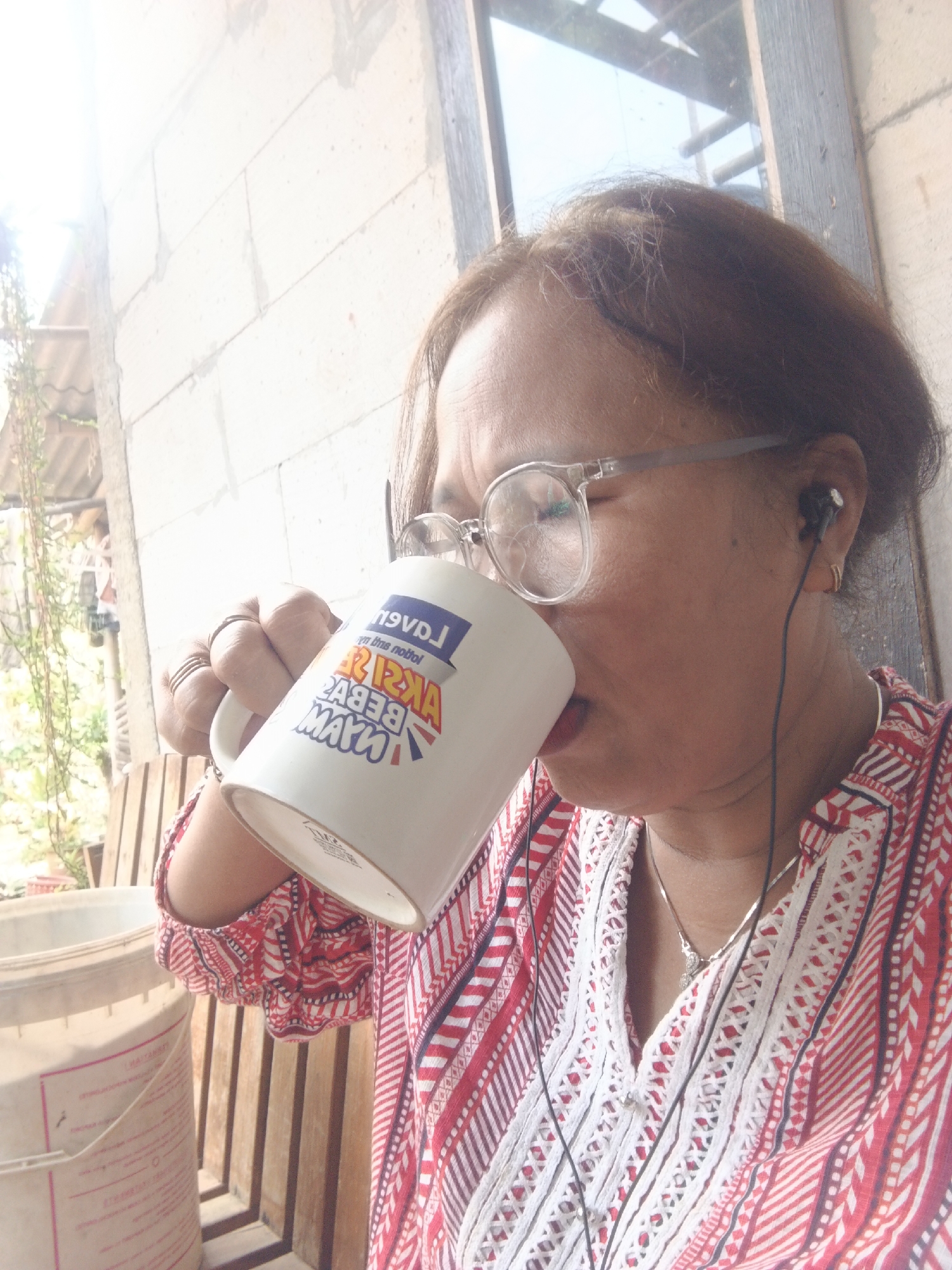 In an outdoor scene in Warungkiara, Indonesia, a woman with medium-length brown hair and glasses drinks from a white mug. She wears a red and white patterned shirt and an earring. A wire earbud is in her right ear. The background shows a white brick wall and a wooden structure. Sunlight illuminates the scene from the left.