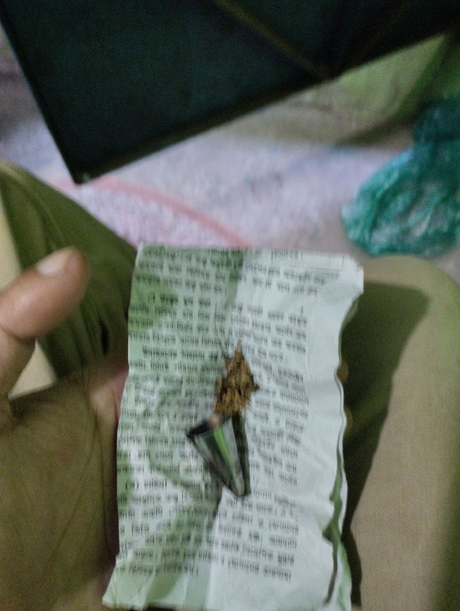 A close-up, dimly lit, and blurry photograph shows a person's thumb and forefinger holding a small piece of white paper with text printed in what appears to be Bengali script. A small, metallic object, possibly a cigarette filter, is attached to the paper, and a piece of brown, dried material is placed on top of it. The background is out of focus, with hints of fabric and a crumpled green plastic bag. The setting is not identifiable as a specific location in Dhaka, Bangladesh, or any other city.