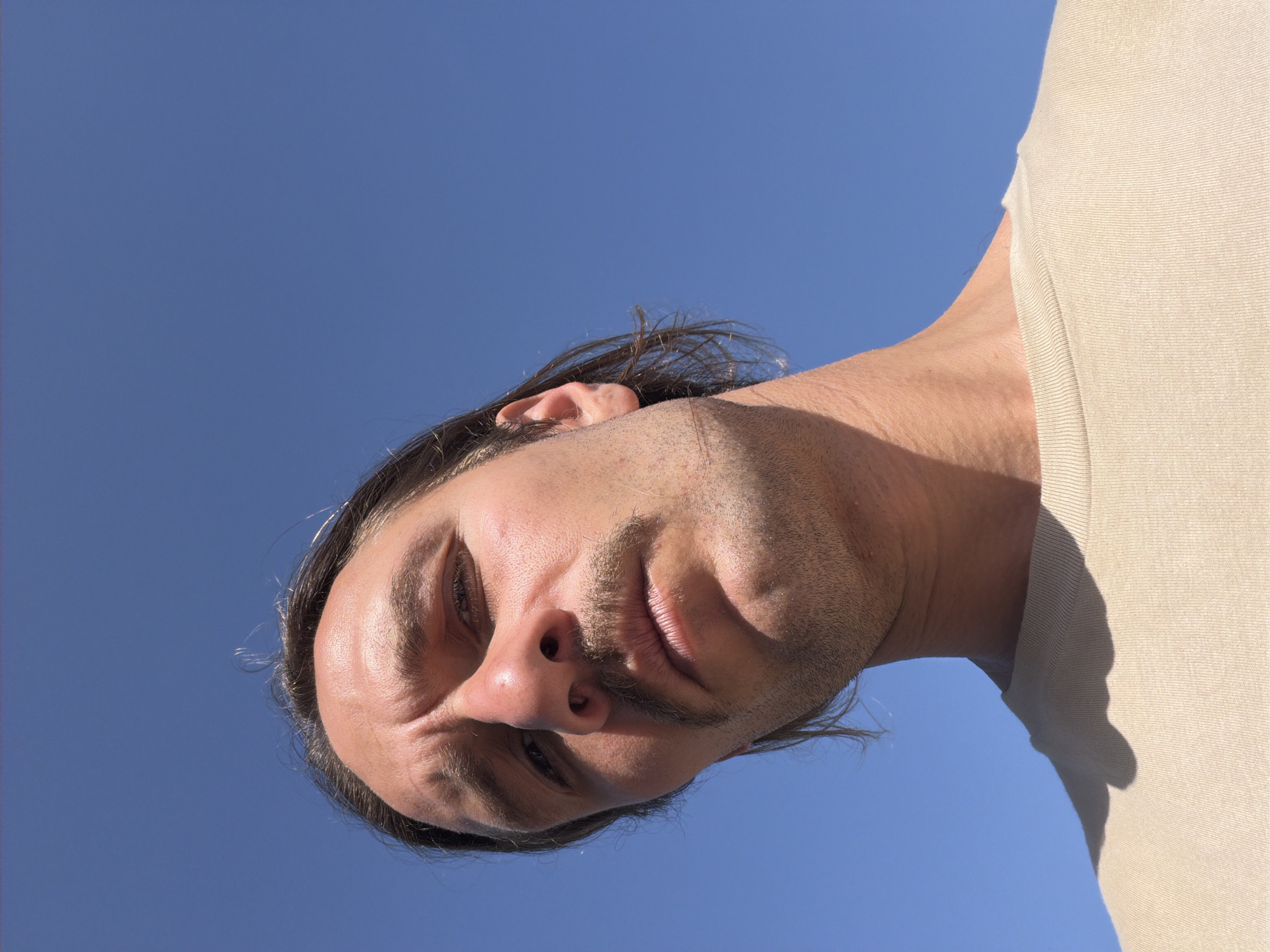 A low-angle, close-up shot depicts a man's face against a clear blue sky. He has dark hair pulled back from his face, fair skin, and a light beard. He is wearing a tan t-shirt. The background is devoid of any landmarks or features suggestive of Petrovac na Moru, Montenegro or any other specific location. The only interaction depicted is the man looking down at the camera. The scene is outdoors and brightly lit.