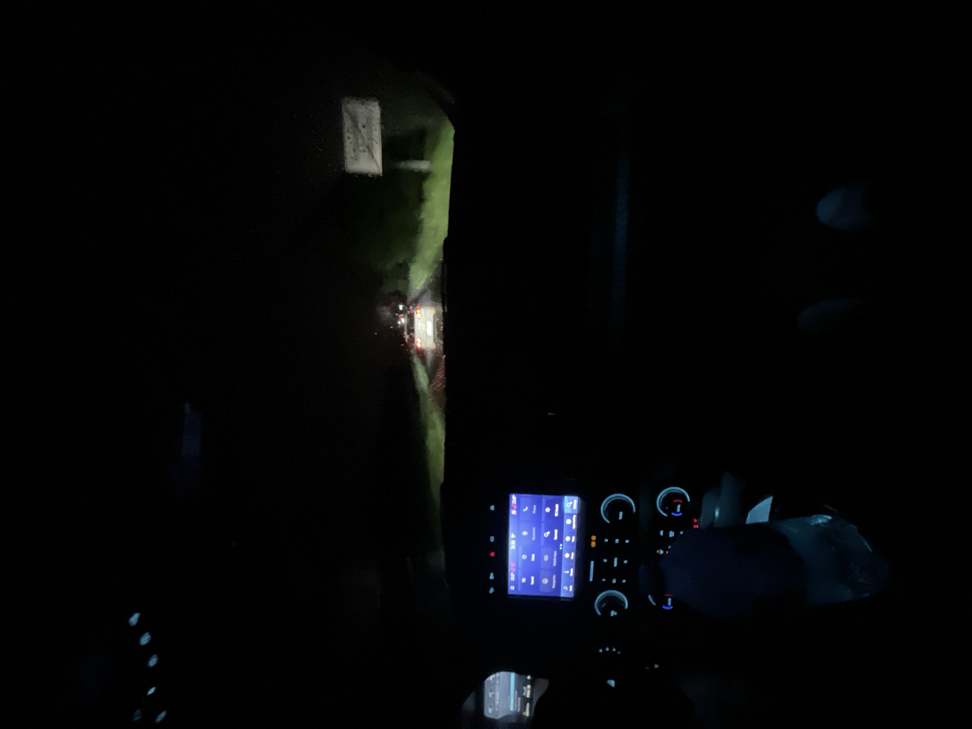 The image depicts a night scene from inside a vehicle. Through the windshield, a road is visible with oncoming headlights and a reflective sign. The interior of the vehicle is dark, but the dashboard is illuminated. It is not possible to determine the specific location in Batanovtsi, Bulgaria, from this image alone. There are no people visible in the frame, only the interior of the vehicle and the view through the windshield.