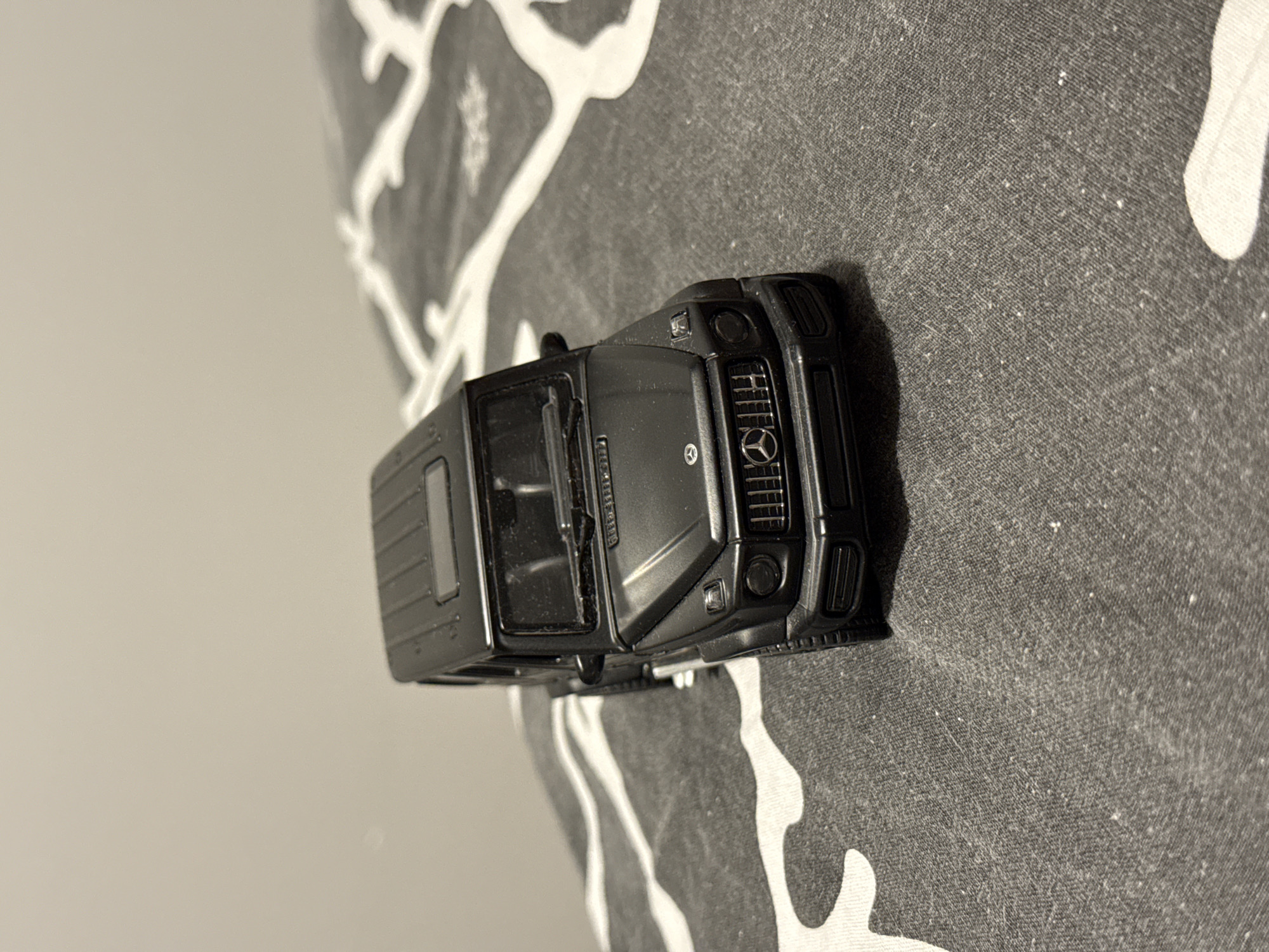 A black toy Mercedes-Benz G-Class is positioned diagonally on a patterned surface. The toy car faces left, with its front grille and headlights clearly visible. The surface beneath the car features a repeating abstract white pattern on a dark grey background. The background of the image is a plain, light grey color. There are no people or any indications of the city of Łódź, Poland present in the image.