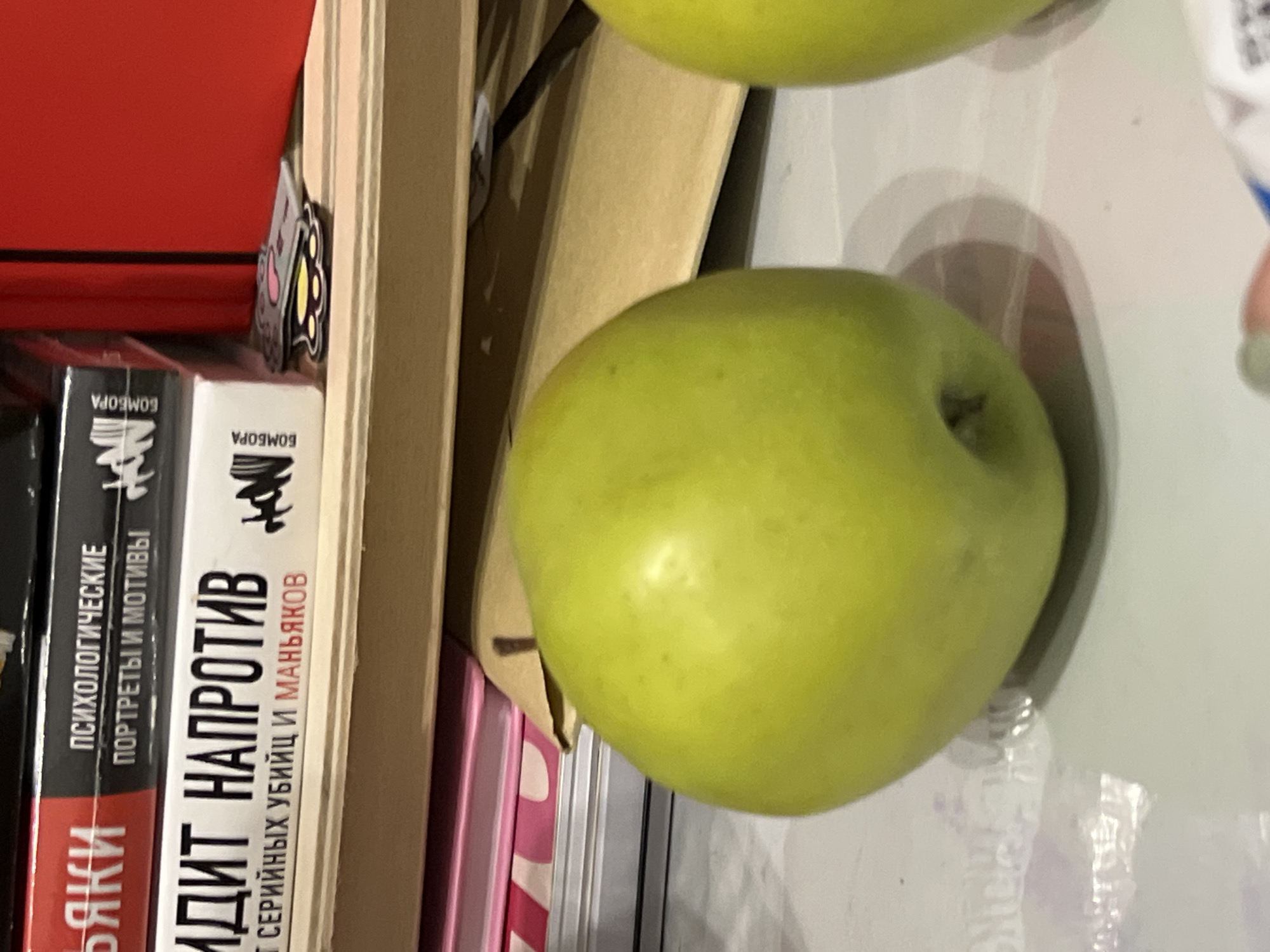 The image is a close-up, eye-level, indoor shot of two green apples on a white, reflective surface. The apple in the foreground is slightly to the left of the frame and is predominantly in focus. Its skin is a vibrant green with some lighter patches and small speckles. The stem is visible in a small indentation. A second green apple is partially visible on the right side of the frame, also out of focus.
Behind the apples, stacked books are visible on shelves. The spines of several books are discernible. One book, in the upper left, has red and black text. Another book below it has white text on a black background and a black and white illustration. The text on these books appears to be in Russian. There is also a glimpse of a red object, possibly another book or binder, to the far right.
A light brown, folded piece of paper or cardboard is positioned between the books and the apples. A small, cartoonish sticker, possibly a cat paw print, is visible on the shelf near the books. The background is cluttered with other items, suggesting a desk or shelf in a room.
The image does not contain any specific landmarks or identifiable features that would indicate its location within the city of Kazan, Russia. The scene is a still life composition focused on the apples and the surrounding objects. There are no people or actions depicted.
