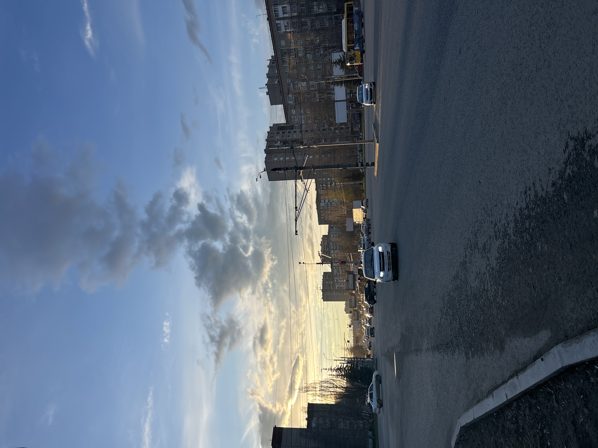 The image captures a wide-angle view of a city street in Izhevsk, Russia, during what appears to be late afternoon or early evening, characterized by a golden hour glow. The sky, transitioning from a clear blue at the top to a radiant yellow-orange near the horizon, is dotted with various clouds, some dark and voluminous, others wispy and illuminated by the low sun.
Along the left side of the frame, several multi-story residential buildings, consistent with Soviet-era architecture, line the street. Their facades are a muted grey or brown, and many windows are visible. Bare tree branches are discernible in front of some buildings. A yellow and blue trolleybus, with indistinct text on its side, is parked or moving slowly next to a building on the left. Numerous utility poles and a complex network of overhead wires, likely for trams or trolleybuses and power, stretch across the street.
The asphalt road occupies the foreground and extends into the distance, appearing wet in places or reflecting the sky. Several cars are visible on the road; one white car with its headlights on is prominent in the middle-right of the frame, and another white car is partially visible further down. A concrete curb marks the edge of the road, with some rough ground or vegetation beyond it. No people are clearly visible in the scene. The overall atmosphere is tranquil, with the warm light lending a serene quality to the urban landscape.