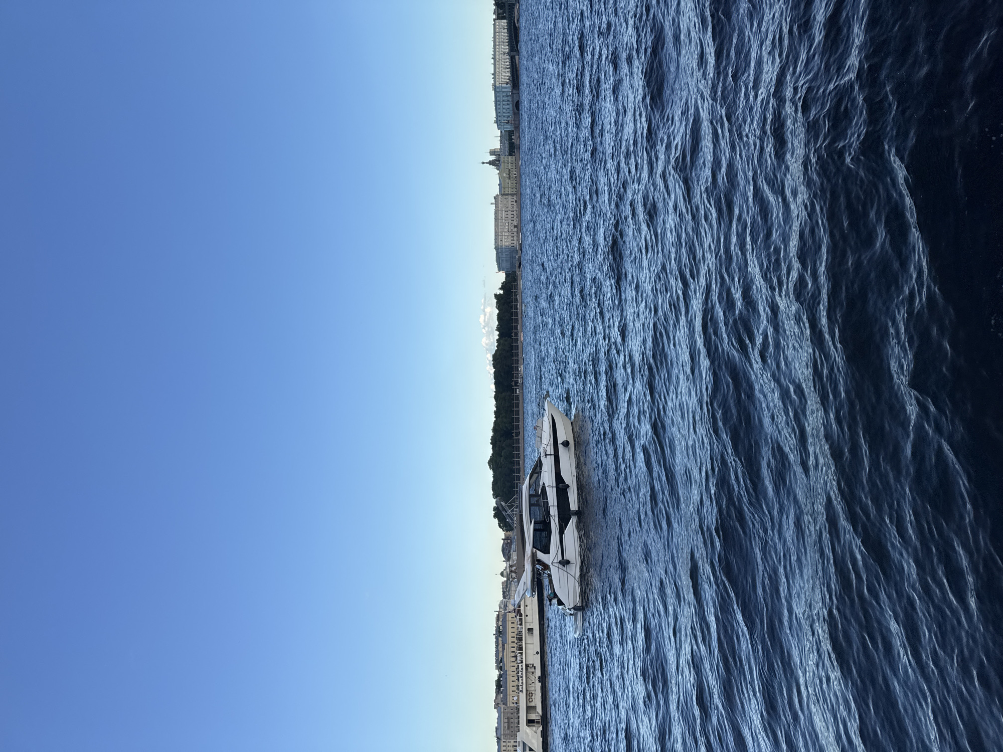 A wide-angle shot captures a modern white yacht with black trim moving across a deep blue, slightly choppy river. The yacht is positioned in the foreground, slightly to the right of the center. In the background, a long, low white boat or ferry travels parallel to the shore. Beyond the boats, a picturesque cityscape of Saint Petersburg, Russia, unfolds. Distinctive architecture with classical facades, domes, and spires are visible, bathed in soft, natural light under a clear blue sky. Green trees line the distant bank, adding a touch of nature to the urban panorama. The water's surface is textured with ripples, indicating gentle movement. The overall scene suggests a serene day on the Neva River.