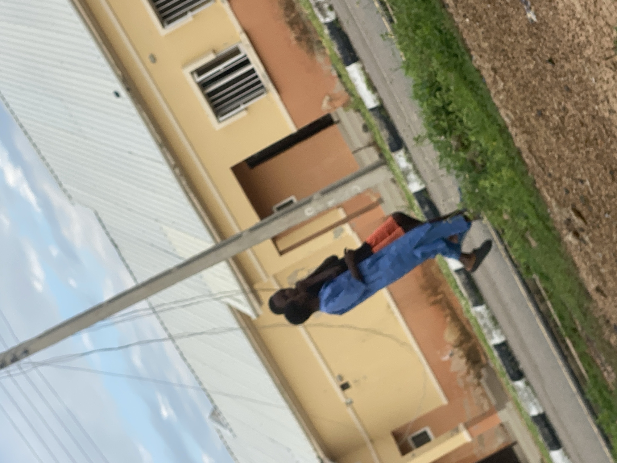 In Dumnga, Nigeria, two individuals are captured in a photograph taken outdoors. A man in a blue garment stands next to another person, partially obscured. They are positioned near a yellow and brown building with a white corrugated roof. A utility pole with wires runs vertically through the frame, angled slightly. The ground in front of the building is paved with a road marked with black and white curbs, bordered by green vegetation and brown soil. The sky above is partly cloudy.