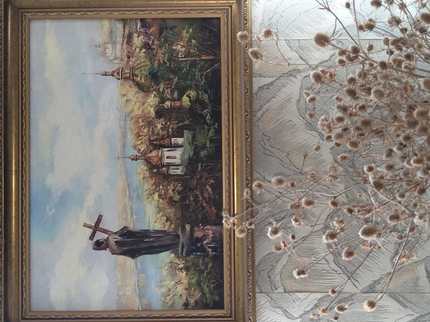 A painting hangs in a gold frame against a textured gray background. The painting depicts a scenic view of a city with a river, churches, and a statue of a figure holding a cross. In the foreground, a bouquet of dried, fluffy plants is arranged in front of the painting.