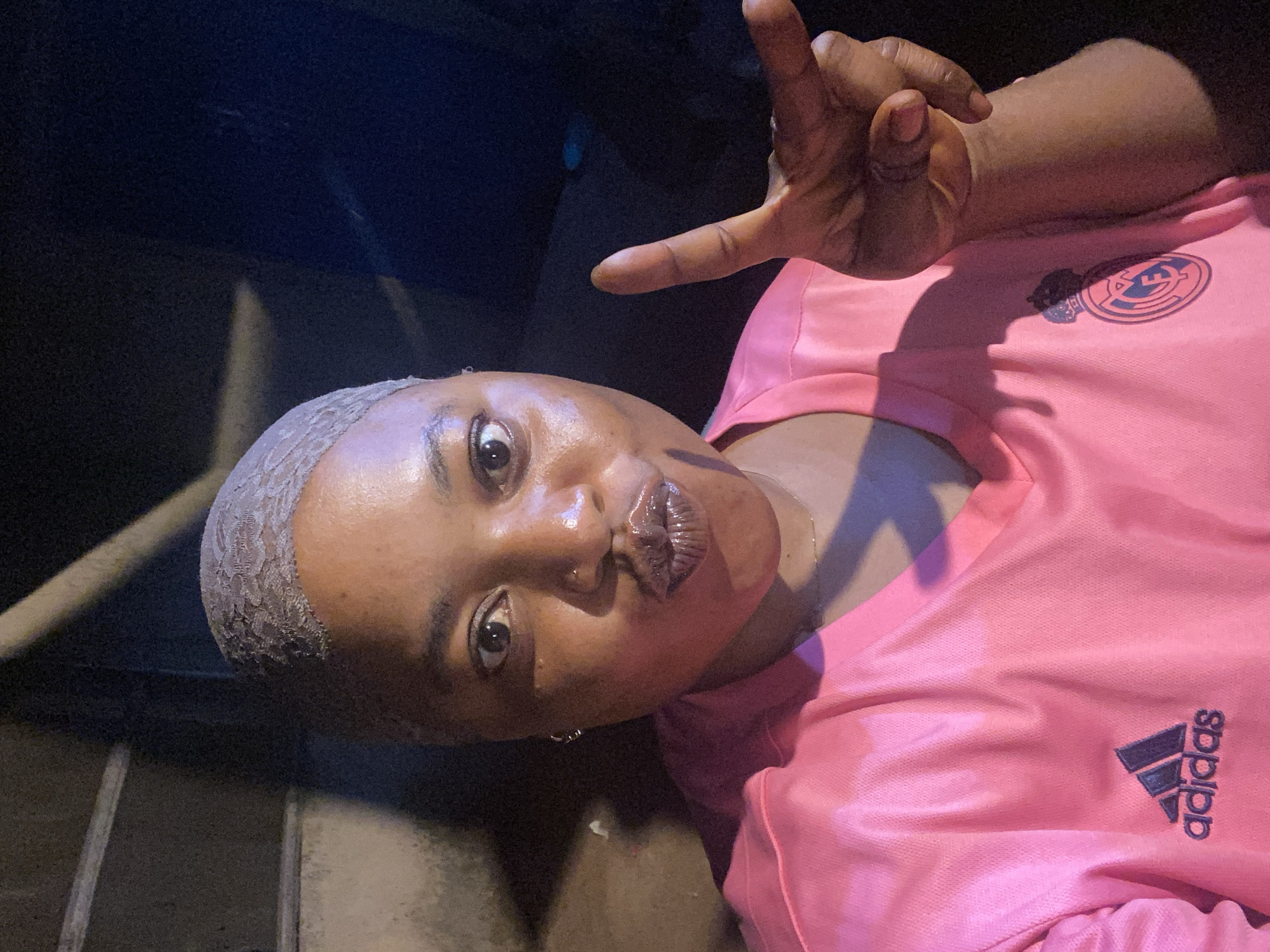 A close-up, front-facing, waist-up shot captures a Black female in Du, Nigeria, wearing a pink Adidas jersey. She has a clear lace frontal wig on her head and is making a kissing face, with her lips pursed. Her right hand is raised, with two fingers extended in a peace sign. The background is dark and indistinct, with what appears to be concrete stairs or a wall to her left. The lighting is dim, creating strong shadows on her face and jersey.