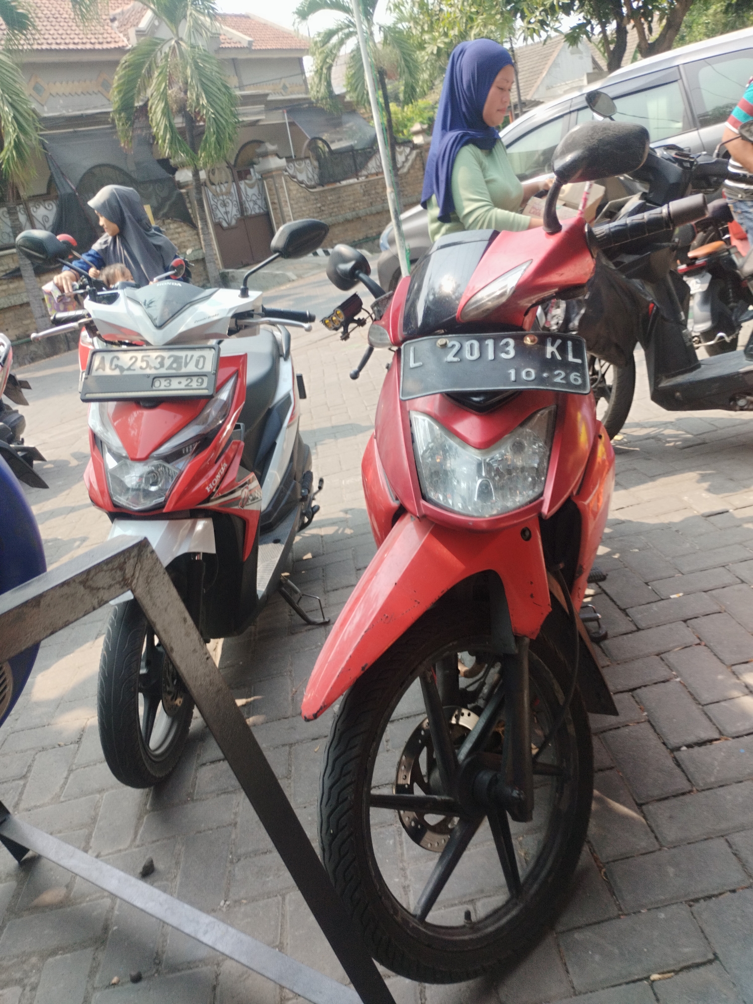 In Surabaya, Indonesia, two red and white scooters are parked on a brick-paved street. The scooter on the left has a license plate reading "AG. 2532 VO" and "03-29". A woman wearing a blue hijab and a light green shirt is partially visible behind the handlebars of this scooter. The scooter on the right, which is entirely red, displays a license plate with "L 2013 KL" and "10-26". In the background, a silver car is parked, and a residential building with a tiled roof is visible. Palm trees and other foliage suggest a tropical environment. The image is captured from a low angle, focusing on the scooters and the street.