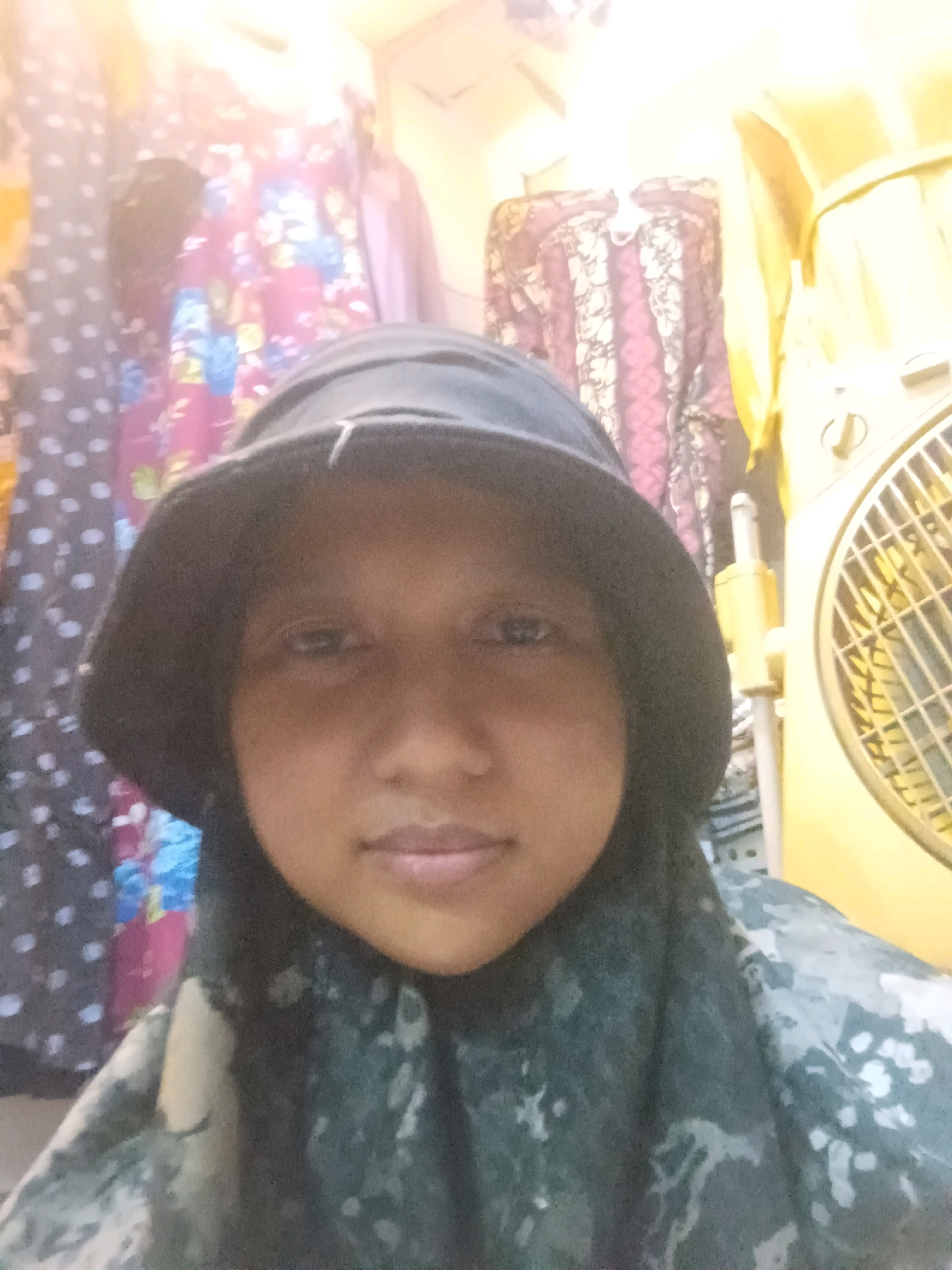 A close-up, selfie-style, medium shot shows a young person wearing a dark bucket hat and a dark, patterned hijab. Their face is centrally positioned, with the camera looking up at them slightly. The lighting is bright and appears to be from above, casting a warm glow on the scene.
In the background, to the left, are vertically arranged fabrics with various patterns. One appears to be a dark gray fabric with white polka dots, and next to it is a fabric with a floral print featuring pink, blue, and green elements. Further to the left, there is a glimpse of a yellow fabric.
To the right of the person, a large, yellow electric fan is visible, with its grille facing forward. Behind the fan and the person, more fabrics can be seen hanging, including a dark red and white patterned textile and a light yellow garment. The overall environment suggests an indoor setting, possibly a shop or a home with stored clothing. The exact location in Sumuragung, Indonesia cannot be determined from the image alone.