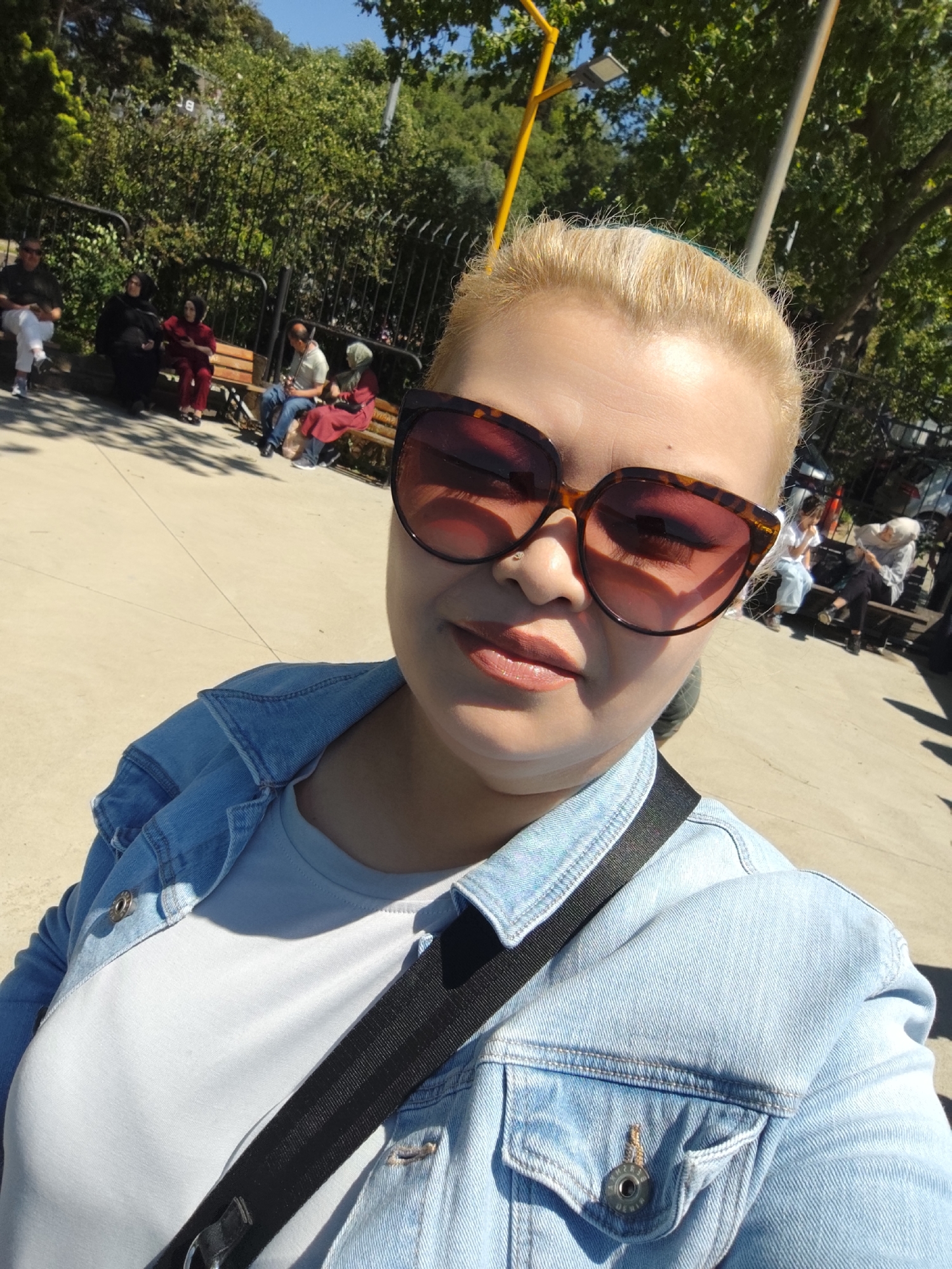 A woman with short blonde hair and large, tortoiseshell-framed sunglasses takes a selfie in a park. She wears a light blue denim jacket over a light gray t-shirt with a black strap across her shoulder. The background features a paved area with trees and several benches occupied by people. The location is Sarıyer, Turkey.