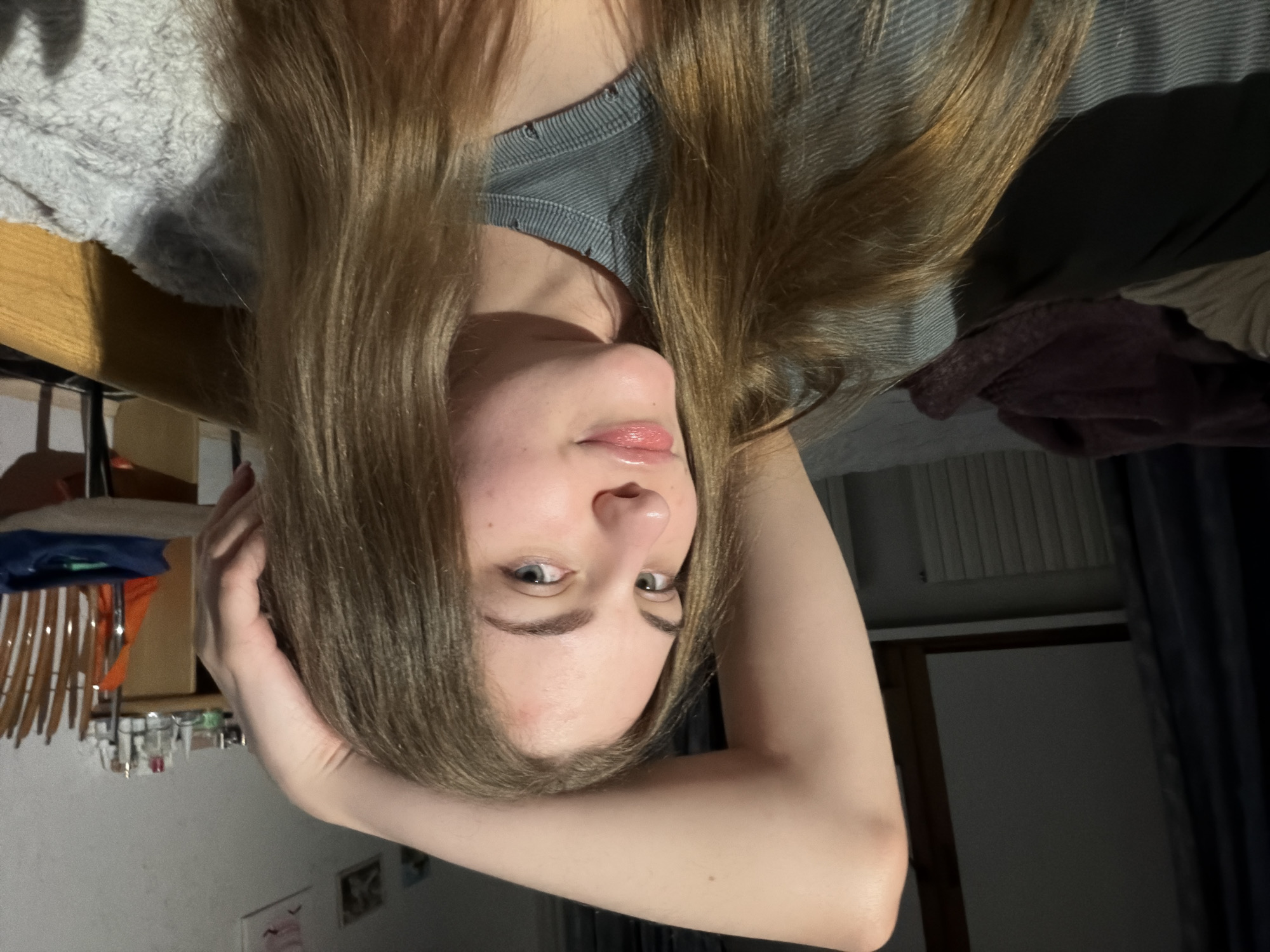 In an indoor, dimly lit bedroom, a young Caucasian woman with blonde hair lies down with her right arm raised behind her head. She is wearing a grey shirt and appears to be looking at the camera. The background shows a bed with blankets, a radiator, a dark curtain, and a wooden dresser with various items on it. A wooden chair with a person's clothing draped over it is also visible. The scene is likely a personal living space in the city of Dnipro, Ukraine.