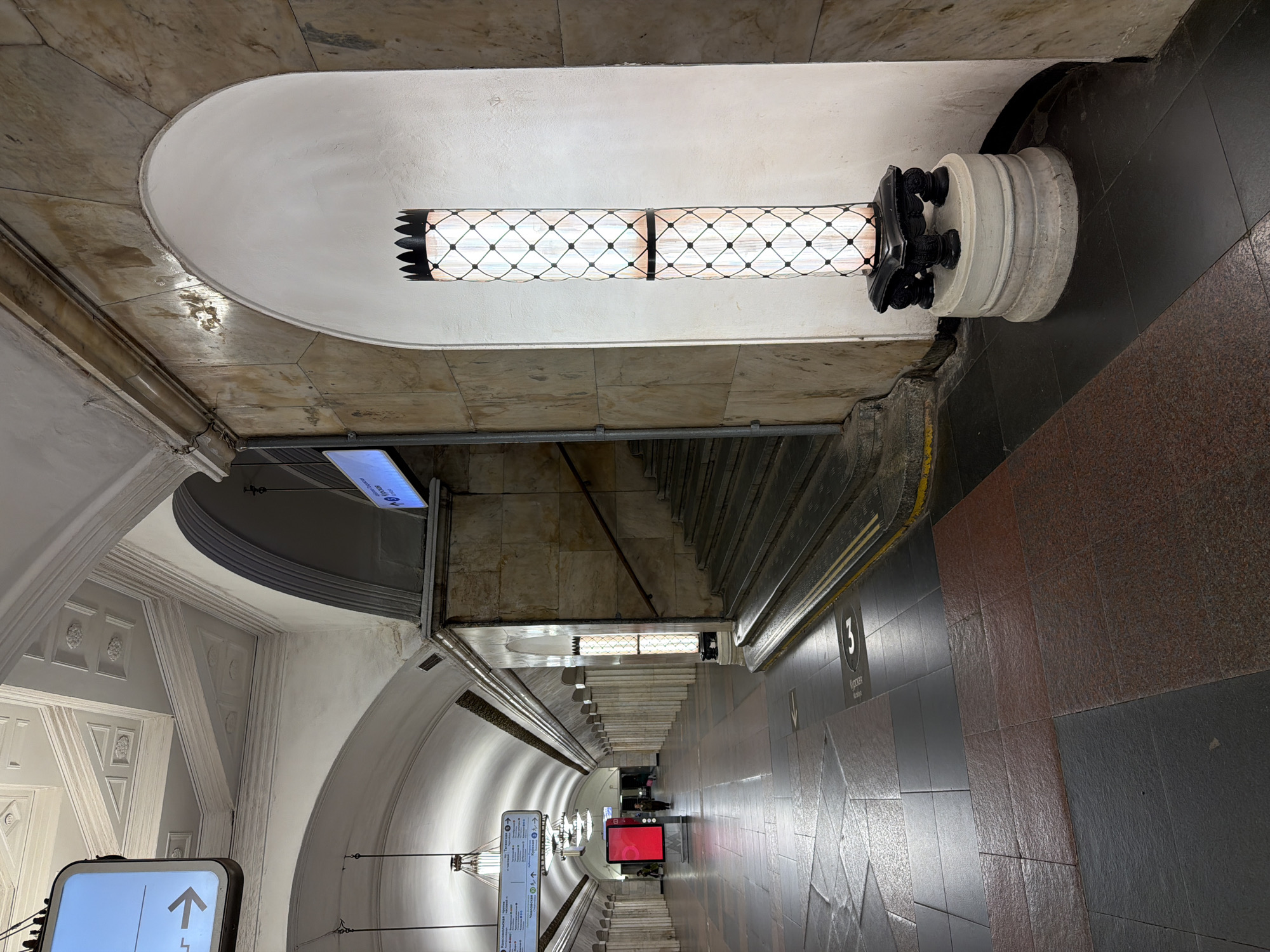 This is an interior view of a Moscow Metro station in Russia. The image is taken from a slightly elevated perspective, looking down the length of a passageway with stairs on either side leading to the platforms.
The architectural design is ornate, featuring arched ceilings, decorative molding, and stone or marble cladding. A cylindrical lamp with a diamond pattern illuminates the space. On the right side of the passageway, there's a tiled wall of brown and black. Signage is visible, including directional arrows and the number "3" with the Cyrillic word "Курская" (Kurskaya), indicating this is the Kurskaya station. A red screen with text is visible farther down the passage. Several people can be seen in the distance.