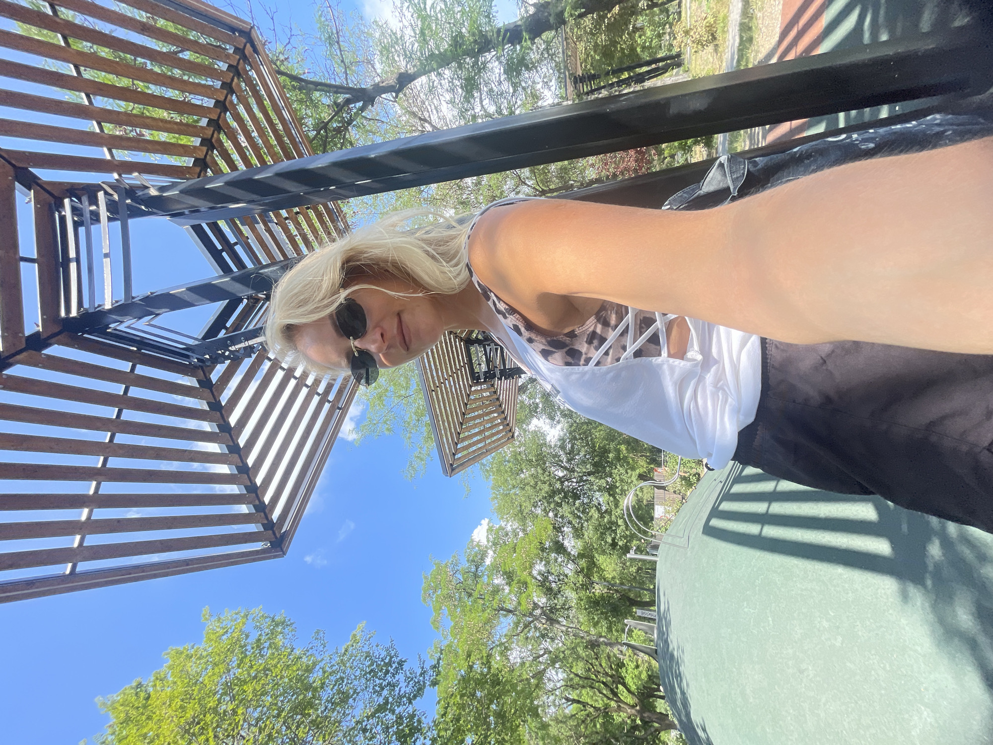 A blonde woman wearing sunglasses, a white tank top over a leopard print bikini top, and black shorts is taking a selfie outdoors. She is standing next to a black metal structure with a wooden slat roof. In the background, there are trees, a blue sky with clouds, and a green, domed surface that appears to be part of a playground. The image was taken at the Novorossiysk Olympic Sports Complex in Novorossiysk, Russia.