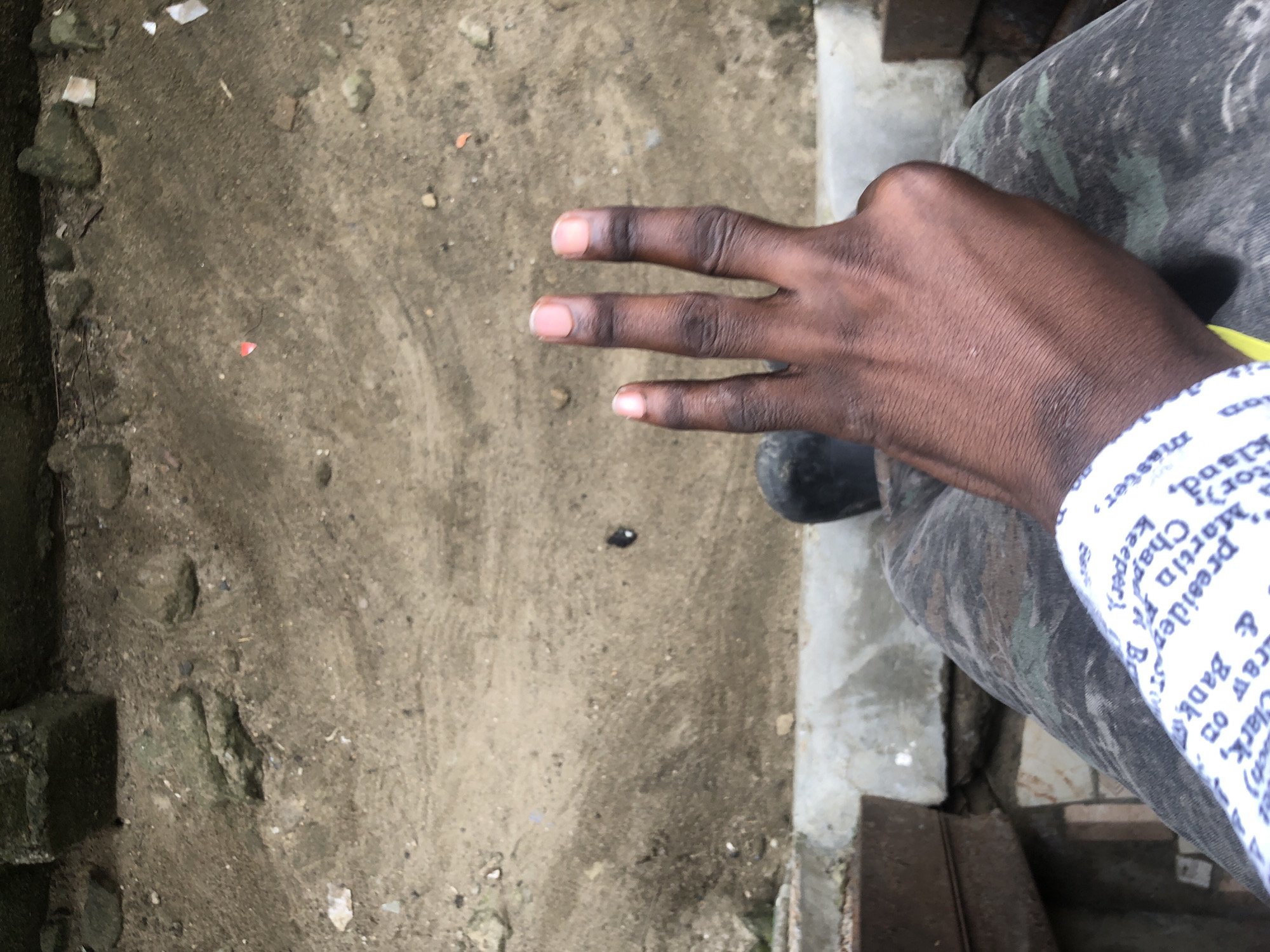 An overhead shot captures a dark-skinned hand with three fingers extended, held up against a background of dry, dusty ground and concrete. The person is wearing camouflage pants and a white shirt with blue text, partially visible on the left side of the frame. The environment appears to be an outdoor urban setting, possibly a courtyard or alleyway, consistent with locations in Ado, Nigeria. The ground is uneven, with scattered rocks and debris.