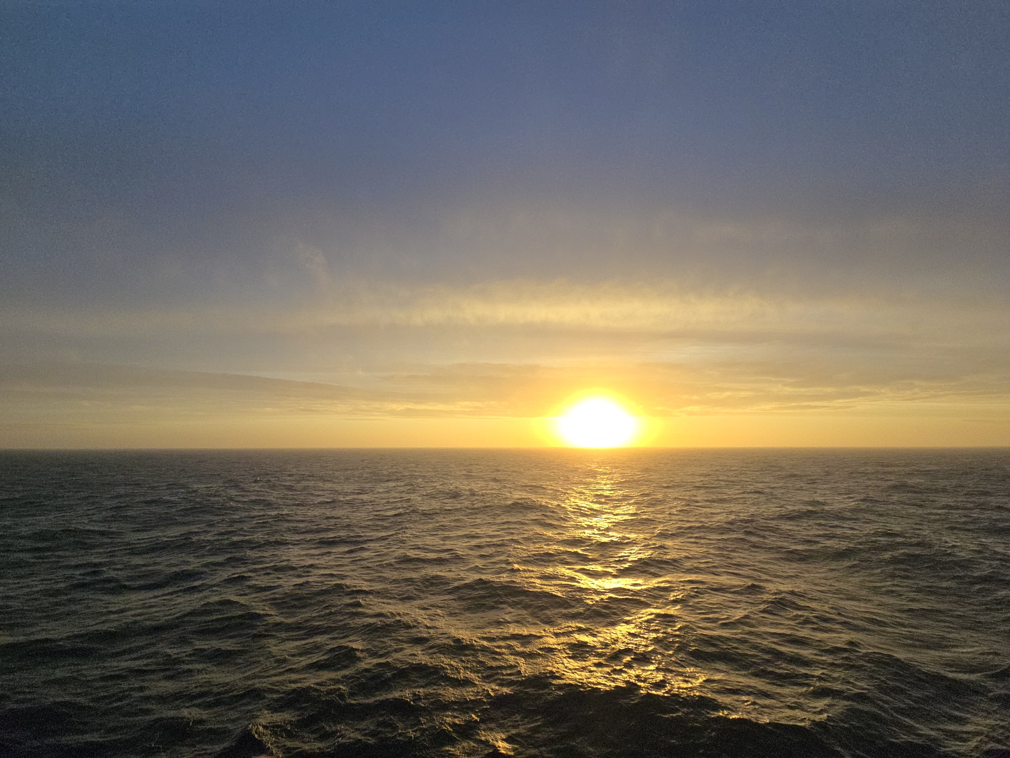 The image depicts a sunset over the ocean. The sun is low on the horizon, creating a bright glow with a reflection on the water. The sky transitions from a dark blue at the top to a lighter, yellowish hue near the horizon. The water is choppy with visible waves. There are no people or objects in the scene other than the ocean and sky. The location is unknown.