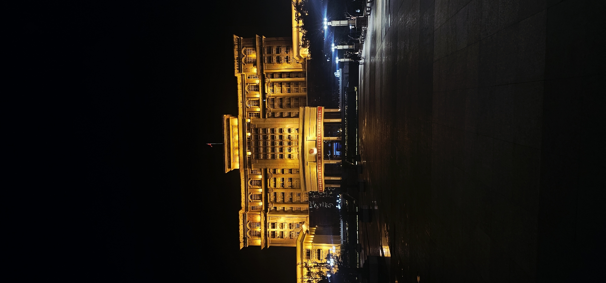 The image shows a well-lit building at night, with warm yellow lights illuminating its facade. The building appears to be a government or official building, with a flag flying from its roof. In front of the building is a large open plaza paved with dark tiles. There are streetlights and some trees visible in the foreground and to the sides. In the distance, two figures can be seen walking across the plaza. The overall scene suggests an urban environment in Beibei, China, likely a public space in front of a significant structure. The wet appearance of the pavement in the foreground might indicate recent rain.