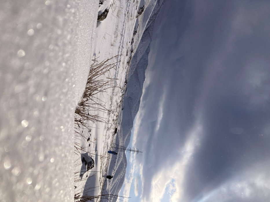 The image depicts a snow-covered mountainous landscape in Churmian, Pakistan, under a partially overcast sky. The foreground features a low-angle perspective of crystalline snow, illuminated by sunlight from the right, creating bright reflections. Scattered dry, brown, leafless bushes and dark rocks interrupt the snow cover. In the mid-ground, rolling snow-covered hills recede into the distance. A large metal lattice electricity pylon stands prominently on a rise to the right of the central frame, with utility lines visible extending across the hills. Two individuals are present in the mid-ground: one wearing a blue outer garment stands to the left of the pylon, and another wearing light-colored clothing walks further to the right. The sky overhead shows dark, heavy clouds dominating the upper left, transitioning to brighter, broken clouds and visible blue sky with a sun flare on the right.