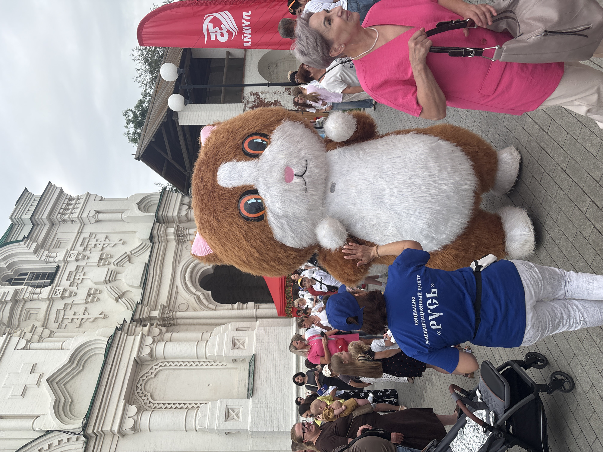 This media file shows an outdoor event with several people and a large mascot costume of a cat. The scene appears to be taking place during the day, under an overcast sky. The background features a white, ornate building with intricate architectural details, suggesting a historical or culturally significant location. A red banner with white Cyrillic text is visible at the top of the image.
The cat mascot is a prominent feature, with brown and white fur, large orange and black eyes, and pink ears and nose. It is interacting with a person wearing a blue t-shirt. The t-shirt has Cyrillic text on the back which reads "СОЦИАЛЬНО-РЕАБИЛИТАЦИОННЫЙ ЦЕНТР «РУСЬ»" (Social Rehabilitation Center "Rus'").
In the foreground and middle ground, a crowd of people are gathered, some standing and some sitting. Several individuals are holding bags or children. One person is pushing a stroller. The overall atmosphere seems to be one of a community event or festival. The people's expressions are not clearly discernible due to the image quality and distance, but the presence of a mascot and a gathering suggests a positive or engaging event. The paving stones indicate an urban or public square setting.