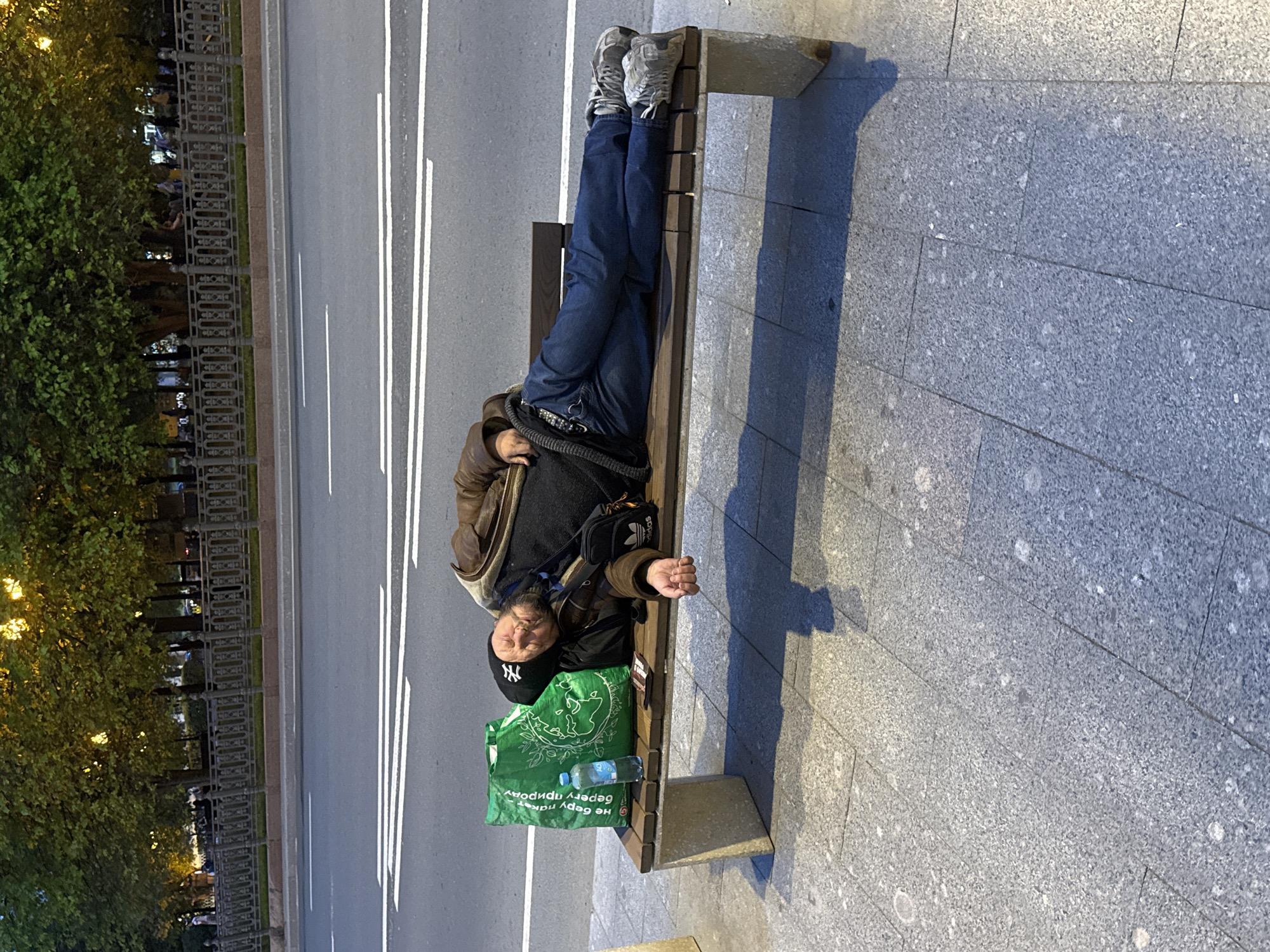 A man is lying on a park bench in Moscow, Russia. He is dressed in jeans, a jacket, and a black baseball cap with the NY logo. He has a green shopping bag next to him. The bench is located next to a street with white lines and a sidewalk made of gray paving stones. Trees and a fence line the other side of the street.