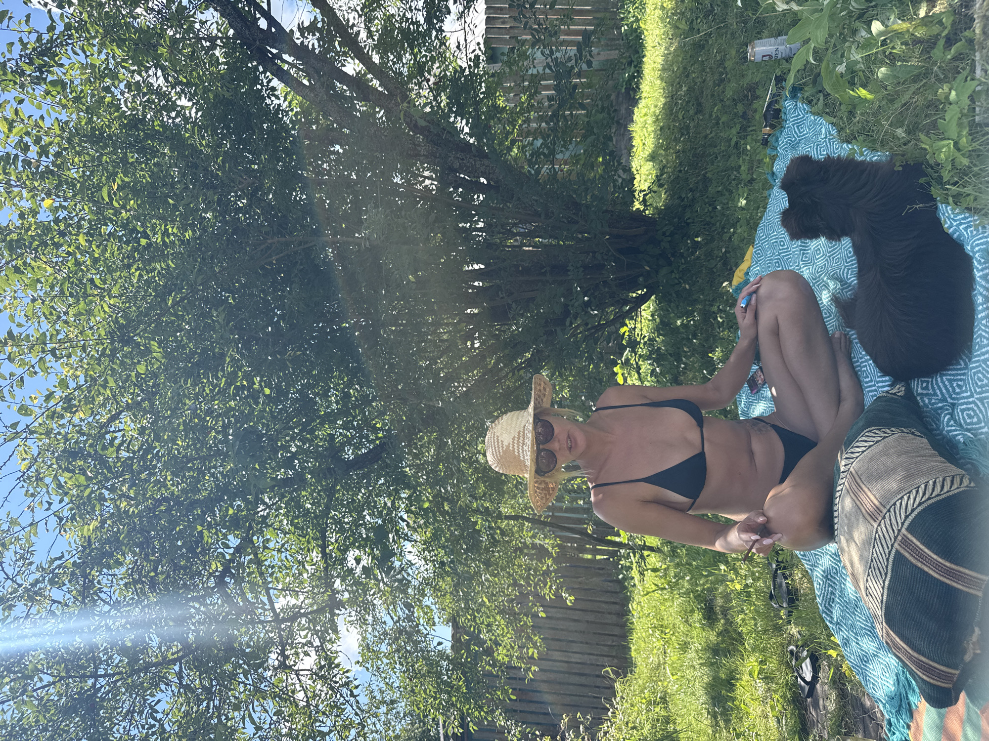 A woman wearing a black bikini and a straw hat is sitting cross-legged on a turquoise blanket in a garden in Красная Пресня Снт, Russia. She is wearing sunglasses and holding a cigarette in her right hand. Next to her is a dark brown dog with fluffy fur. The background shows a wooden fence, green grass, and a large tree with sunlight filtering through its leaves.