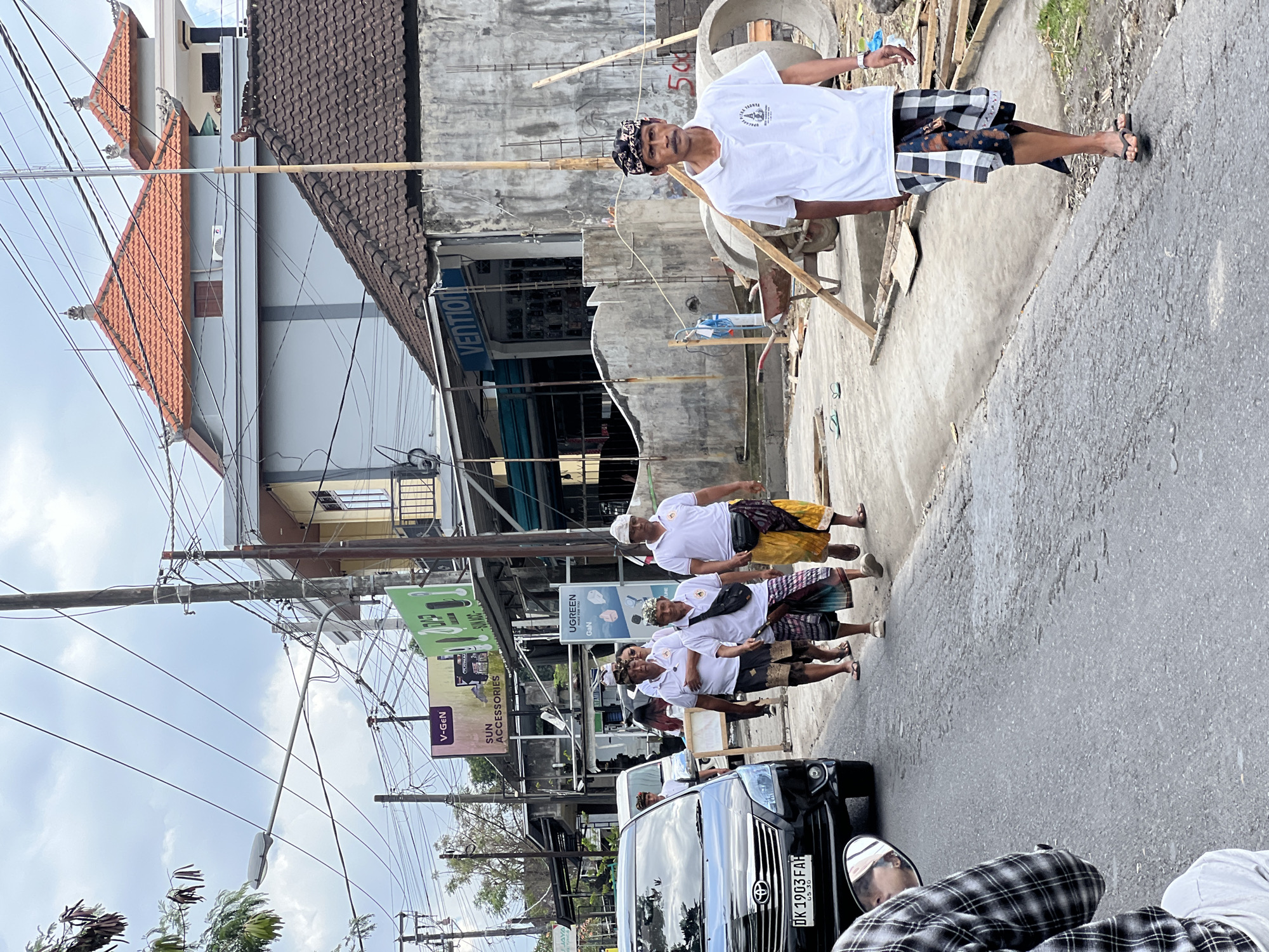 In the city of Ubud District, Indonesia, a group of men are walking along a paved road. The men are wearing traditional Balinese attire, including sarongs and white shirts. One man in the foreground is wearing a white t-shirt with a logo and a black and white checkered sarong. He is also wearing a black headband and flip-flops. Another man is wearing a yellow sarong and a white shirt. The men appear to be walking in the same direction. There is a black car on the left side of the road. In the background, there are buildings and power lines. Construction materials, including concrete pipes and wooden planks, are visible on the right side of the road. The sky is overcast.
