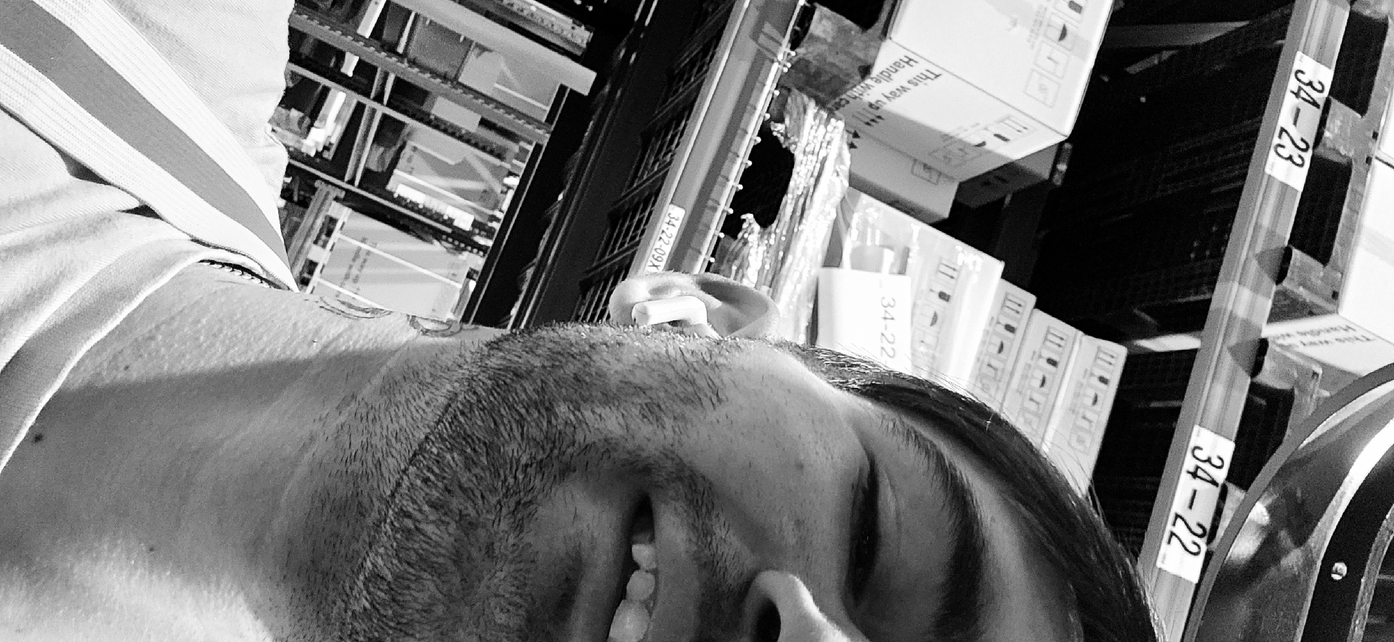 A black and white image shows a man lying on his side with his head tilted back, suggesting he is looking up. The visible part of his body includes his face, neck, and the top of his shoulder, on which a collared garment with striped fabric is seen. He has short hair and a beard. The environment surrounding him seems to be a storage area or warehouse. Shelves are filled with boxes and items, with labels visible on some of the shelves. One label reads "34-22" and another "34-23". The image is rotated, and details of the location, specifically in Plovdiv, Bulgaria, are not discernible from this image.