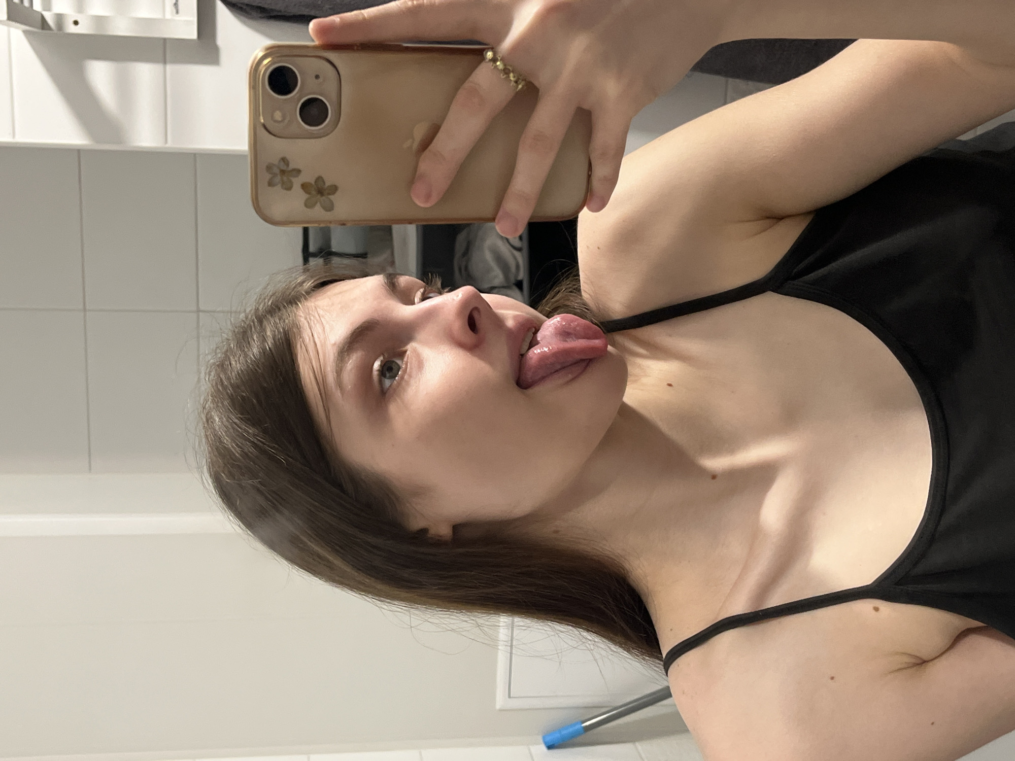 A young woman takes a selfie in a bathroom. She is wearing a black tank top and holding a gold-colored smartphone in her right hand. Her tongue is extended. The background shows a tiled wall and a section of a shelf. There's no indication this image was taken in Saint Petersburg, Russia.