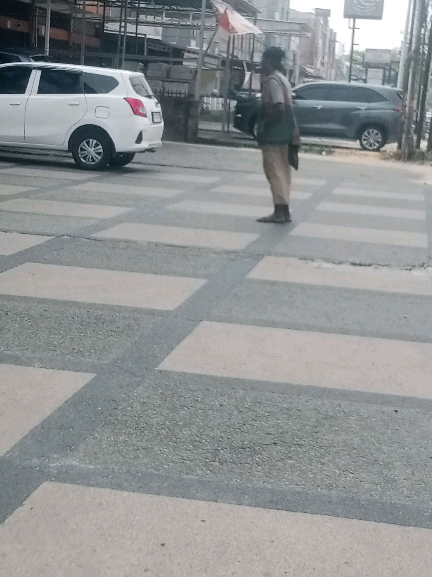 The image shows a person standing on a pedestrian crossing. The person appears to be a man, wearing light-colored pants and a greenish vest over a shirt. He is looking to his left. In the background, there is a white car on the left and a dark SUV on the right, both parked. The pedestrian crossing is marked with white and gray rectangular tiles on an asphalt surface. The scene suggests an urban environment, possibly a street or intersection, within the city of Pekanbaru, Indonesia, given the context provided. The overall lighting indicates it is daytime.