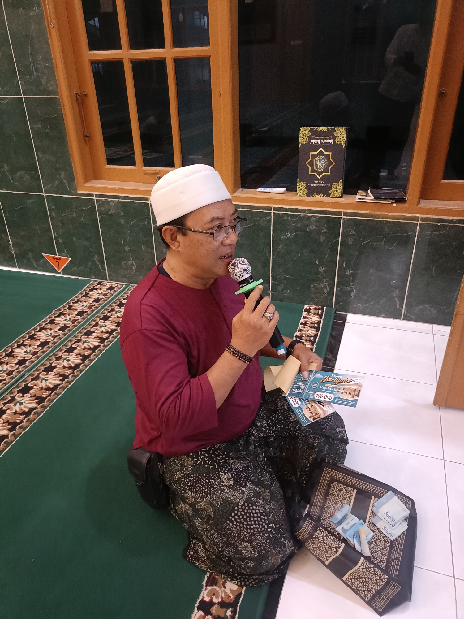 A man wearing a white skullcap and glasses, a maroon long-sleeved shirt, and a patterned sarong sits cross-legged on a green carpet with a decorative border. He is holding a microphone in his right hand and speaking into it. In his left hand, he holds several coupons that appear to be for a mosque donation, each with the text "Kupon Jariyah" and "Masjid As-Sakina" along with a denomination of "100.000". To his left, on the carpet, is a dark, patterned fabric laid out like a prayer mat, with several Indonesian Rupiah banknotes on it, including denominations of 50,000 and 20,000. Behind him, there is a tiled green wall and a wooden-framed window with dark glass, through which a reflection of another person is visible. The scene appears to be indoors, possibly in a mosque or community hall. The specific location of Kediri City, Indonesia, is not visually evident in the image itself but has been provided as context.