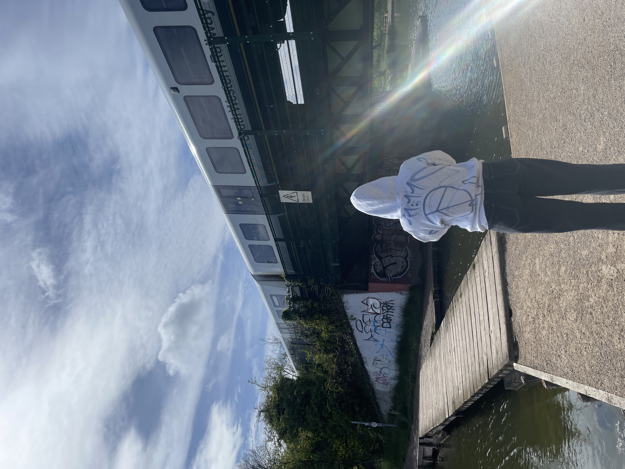 The image captures an outdoor scene featuring a person standing on a paved pathway next to a body of water, with a railway bridge and train overhead. The perspective is from behind the individual, who is wearing a white hoodie with a prominent peace sign and other dark graffiti-style designs on the back, paired with dark trousers.
To the person's right is a calm body of water, likely a river or canal, reflecting the sunlight. A wooden boardwalk or jetty extends along the water's edge. The pathway they are standing on appears to be made of gravel or small stones.
Above and slightly to the left, a railway bridge spans across the scene. A white train carriage, belonging to "South Western Railway," is visible on the bridge. The bridge structure is made of metal girders and includes fencing. Underneath the bridge, concrete walls are covered with various, largely illegible graffiti tags. Dense green foliage and bushes grow along the bank beneath the bridge.
A yellow warning sign featuring an electrical hazard symbol (lightning bolt in a triangle) and the word "Danger" is affixed to the bridge structure.
The sky is bright and blue with scattered white clouds, indicating a clear, sunny day. A strong sun flare or lens flare with rainbow hues emanates from the upper right portion of the image, suggesting the sun is shining brightly from that direction. The overall impression is a vibrant, sunny day in an urban setting, possibly along a waterfront path in Portsmouth, United Kingdom.
