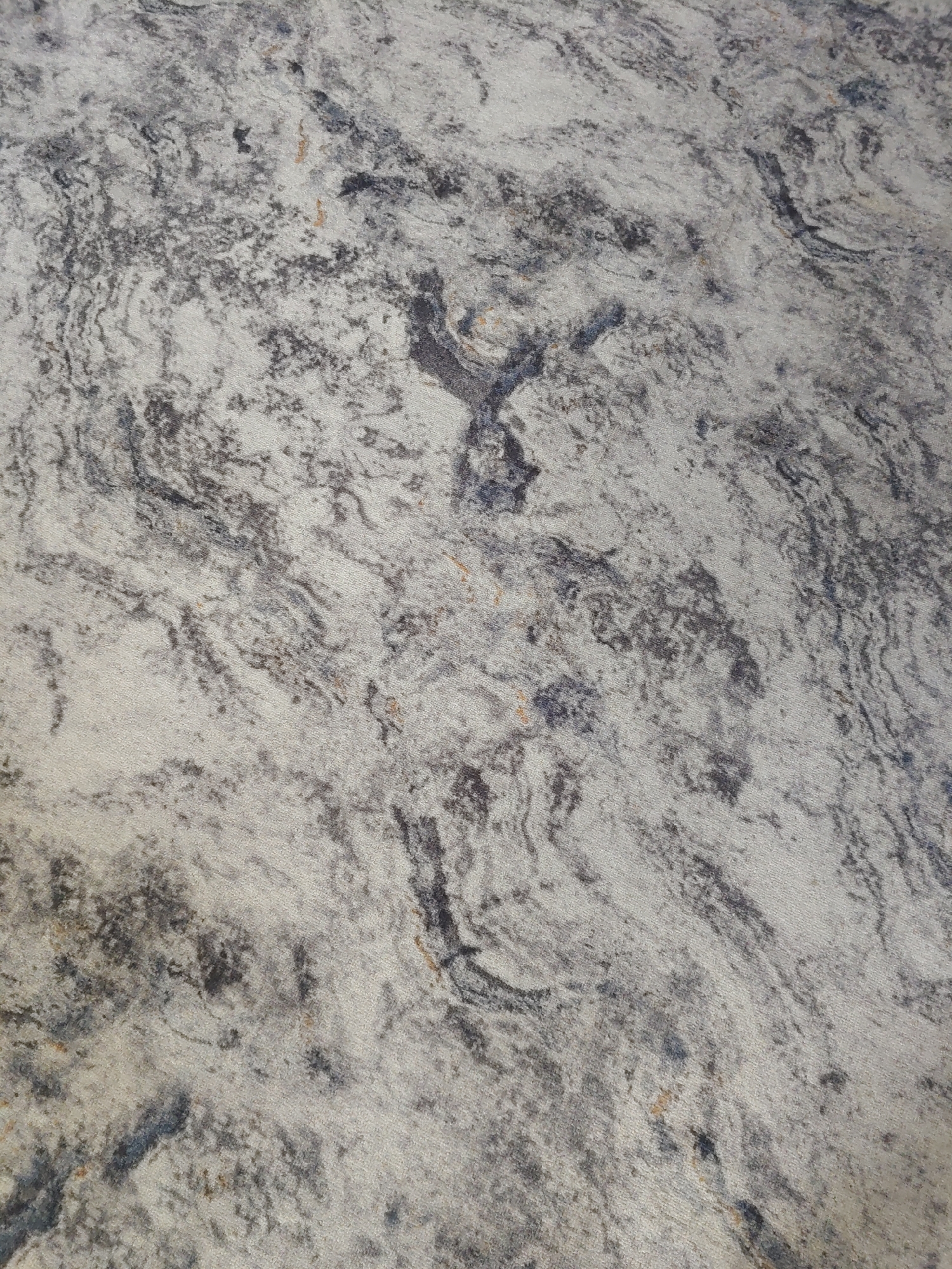 This is a close-up, high-angle, full-frame shot of a textured surface that appears to be a carpet or rug. The pattern consists of swirling veins of dark gray and black against a lighter, off-white or pale gray background. There are also subtle hints of brown and blue within the darker areas. The texture of the fibers is visible, creating a slightly uneven and soft appearance. There are no people, objects, or environmental features visible that would indicate a specific location within the city of Курашим, Russia. The image exclusively depicts the abstract pattern of the material.