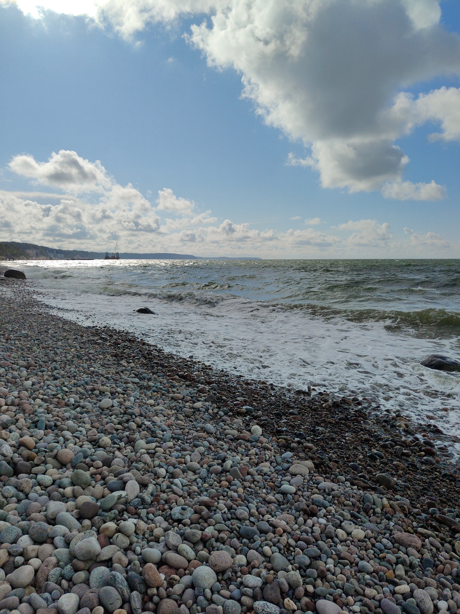 The image shows a pebble beach in Pionersky, Russia. The beach is covered in a multitude of smooth, rounded pebbles of various sizes and colors, ranging from light grey and white to darker shades of brown and black. The pebbles are densely packed, creating a textured foreground.
The sea is visible in the background, with moderately rough, choppy waves breaking onto the shore, creating white foam. The water appears to be a greenish-grey color. In the distance, a hazy coastline is visible, with what appears to be a town or settlement with buildings and possibly some industrial structures or cranes. A few sailboats or masts are also discernible on the horizon.
The sky is partly cloudy, with a bright blue sky peeking through between large, fluffy white and grey clouds. Sunlight is casting some glare on the water, suggesting a sunny day with some wind. There are no people or animals visible in the image. The overall impression is of a natural, somewhat windswept coastal scene.