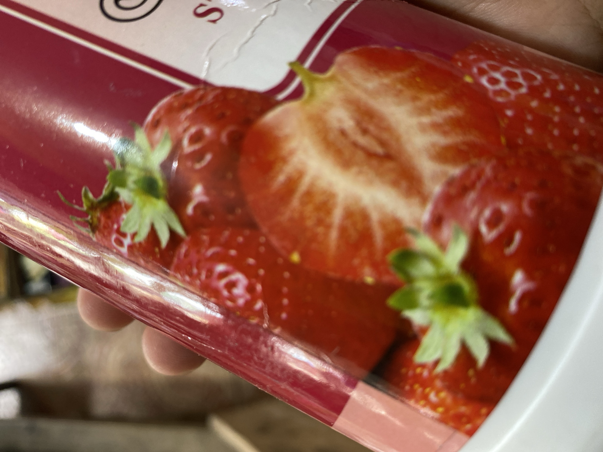A hand is holding a cylindrical product with a label featuring multiple images of strawberries. One strawberry is shown whole with its green stem and leaves, while another is cut in half, revealing its textured interior. The label has a red background with some text in white and a pink border at the bottom. The product appears to be in a retail setting, possibly a supermarket or store, but no specific location within Ta', Indonesia can be determined from the image. The lighting suggests an indoor environment.