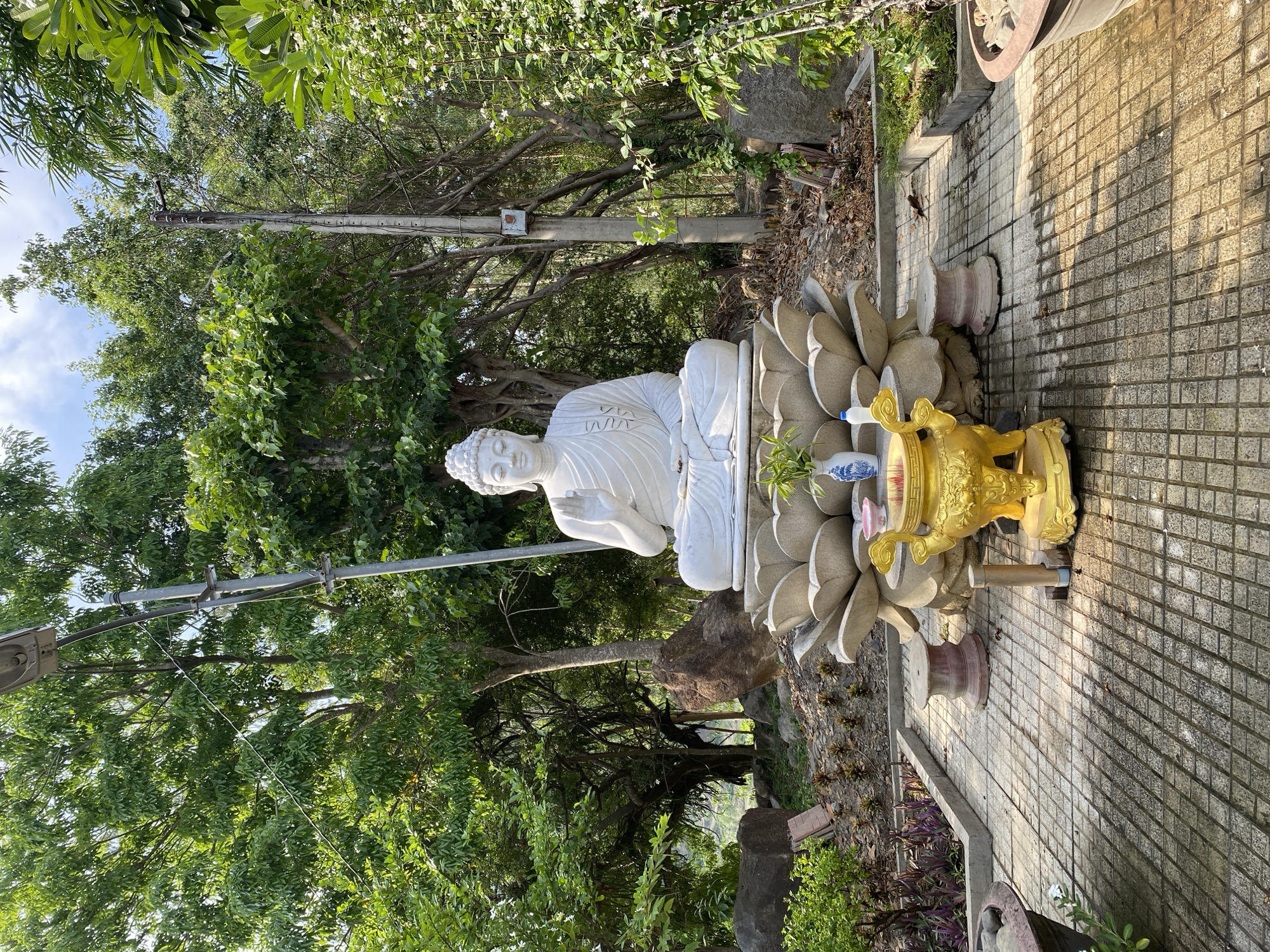 The image depicts an outdoor scene with a large white Buddha statue as the central subject. The statue is seated on a lotus-shaped pedestal, which rests on a paved area made of square tiles. In front of the statue, on the tiled ground, is a golden incense burner with ornate carvings. To the left of the incense burner, there are two decorative vases.
The background is filled with lush green trees and foliage, suggesting a natural or garden setting. A tall utility pole with wires is visible behind the Buddha statue and to its right. The ground in front of the paved area appears to be covered with small stones and some scattered leaves. The lighting suggests it is daytime, with sunlight casting shadows on the tiled ground. The scene is located in Tay Nha Trang Ward, Vietnam.