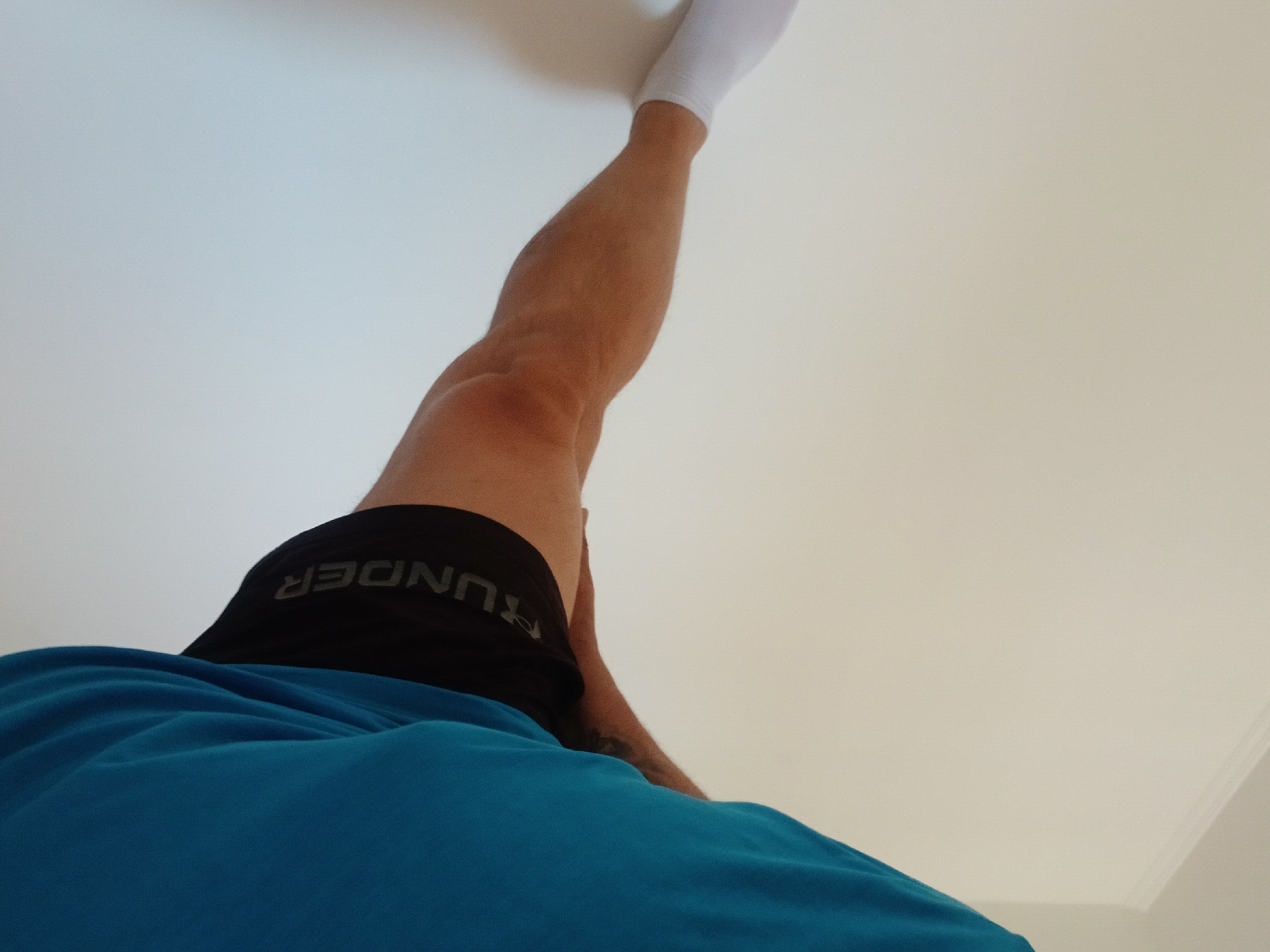 A person's leg, wearing a white sock and black shorts with the word "UNDER" visible, is extended to the left, resting against a white wall. A blue shirt is visible on the right side of the image. The scene appears to be indoors, with the lighting suggesting a daytime setting. There are no discernible landmarks or features that would indicate a specific location within Pfarrkirchen, Germany.