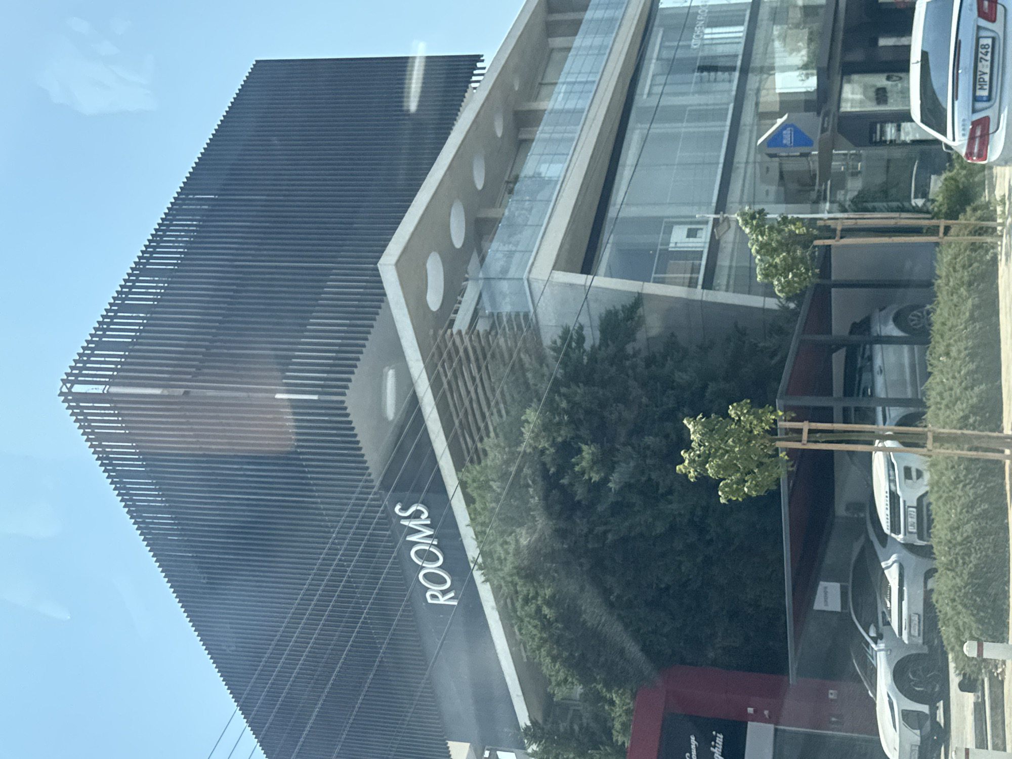 The image shows a modern building with the word "ROOMS" in large white letters on its facade. The building appears to be multi-story with a glass front and a dark, slatted exterior on the upper levels. Trees and shrubbery are visible in front of the building, as well as several parked cars. In the foreground, a silver Mercedes-Benz with license plate "MPV-748" is visible. A red building with the word "Lounge" and text that appears to be "ghini" is to the left of the main building. The scene is consistent with a commercial or hospitality establishment in an urban or suburban setting. The location is Agios Athanasios Municipality, Cyprus.