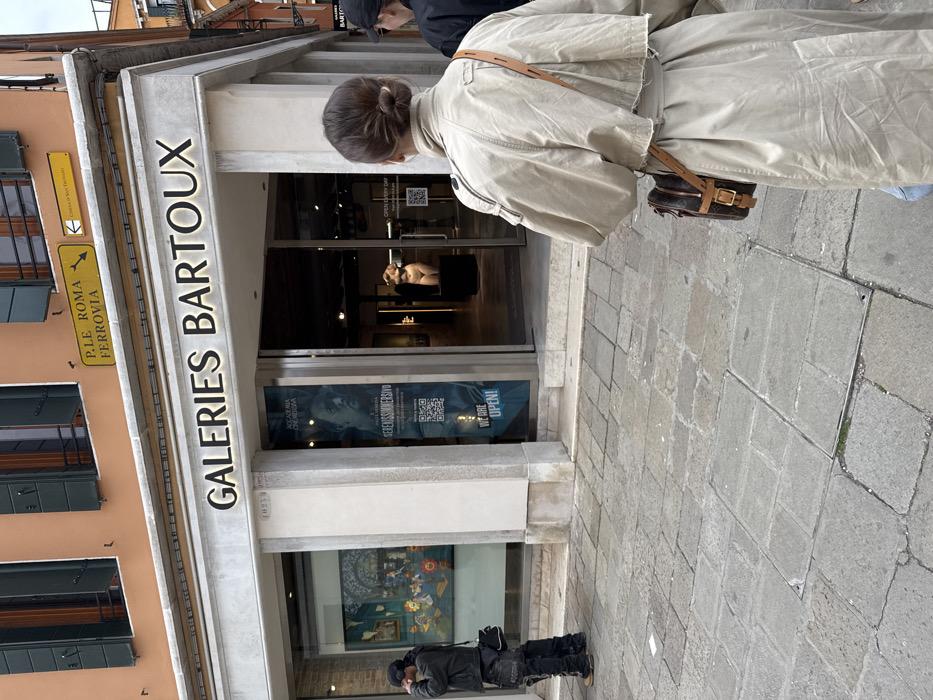 An outdoor, eye-level street view captures a specific location in Venice, Italy, identified by a yellow street sign indicating "P.LE ROMA FERROVIA" (Piazzale Roma Railway Station) with a left arrow, and another sign pointing to "CALLE DI SAN TROVASO." The focal point is the "GALERIES BARTOUX" art gallery, situated on the ground floor of a two-story building with an orange-peach upper facade and a light-colored ground floor. The gallery entrance features large glass doors and display windows.
Inside the left display window, a colorful abstract painting is visible. Through the central glass entrance, a prominent white, nude, full-figured female sculpture rests on a dark pedestal. A blue banner poster affixed to the right door advertises "ACCADEMIA ONLINE VERONA" and "SERENASSIMMERSIVO," including a QR code and the text "WE ARE OPEN!" The building's facade bears the number "1233" near the left side of the gallery.
The scene includes three individuals on the irregularly paved stone walkway. In the immediate foreground, a person is viewed from behind, wearing a light beige trench coat and a brown leather cross-body bag with a distinct monogram pattern. Their dark hair is styled in a bun. To the left, a second person, wearing a dark jacket, dark pants with white lettering, and a dark cap, stands facing right, with their right hand raised towards their head, possibly interacting with a device. This individual carries a small black shoulder bag. Partially visible behind the trench coat wearer, on the far right, is a third individual's head and shoulder, also wearing a dark cap. No direct interaction between the depicted individuals is evident.